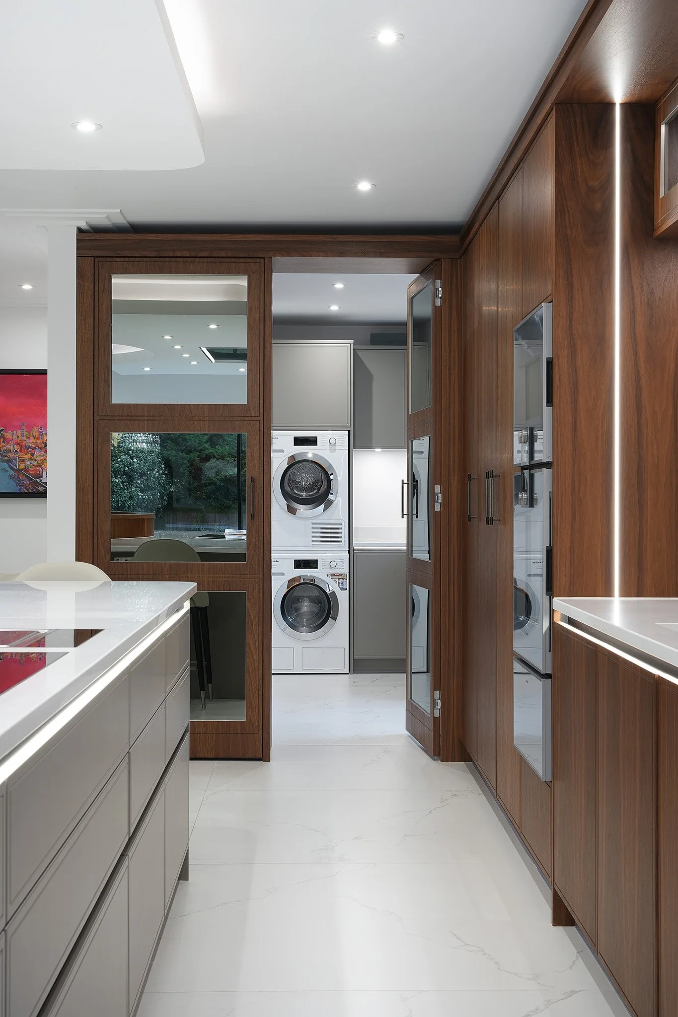 Modern laundry room with wooden cabinets and white appliances, including stacked washer and dryer, visible through a glass door.