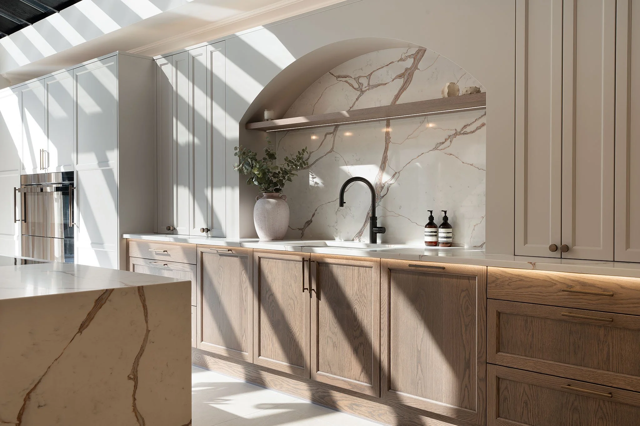 Modern kitchen with cream-colored and wooden cabinets, marble backsplash, black faucet, and decorative items, illuminated by natural light.