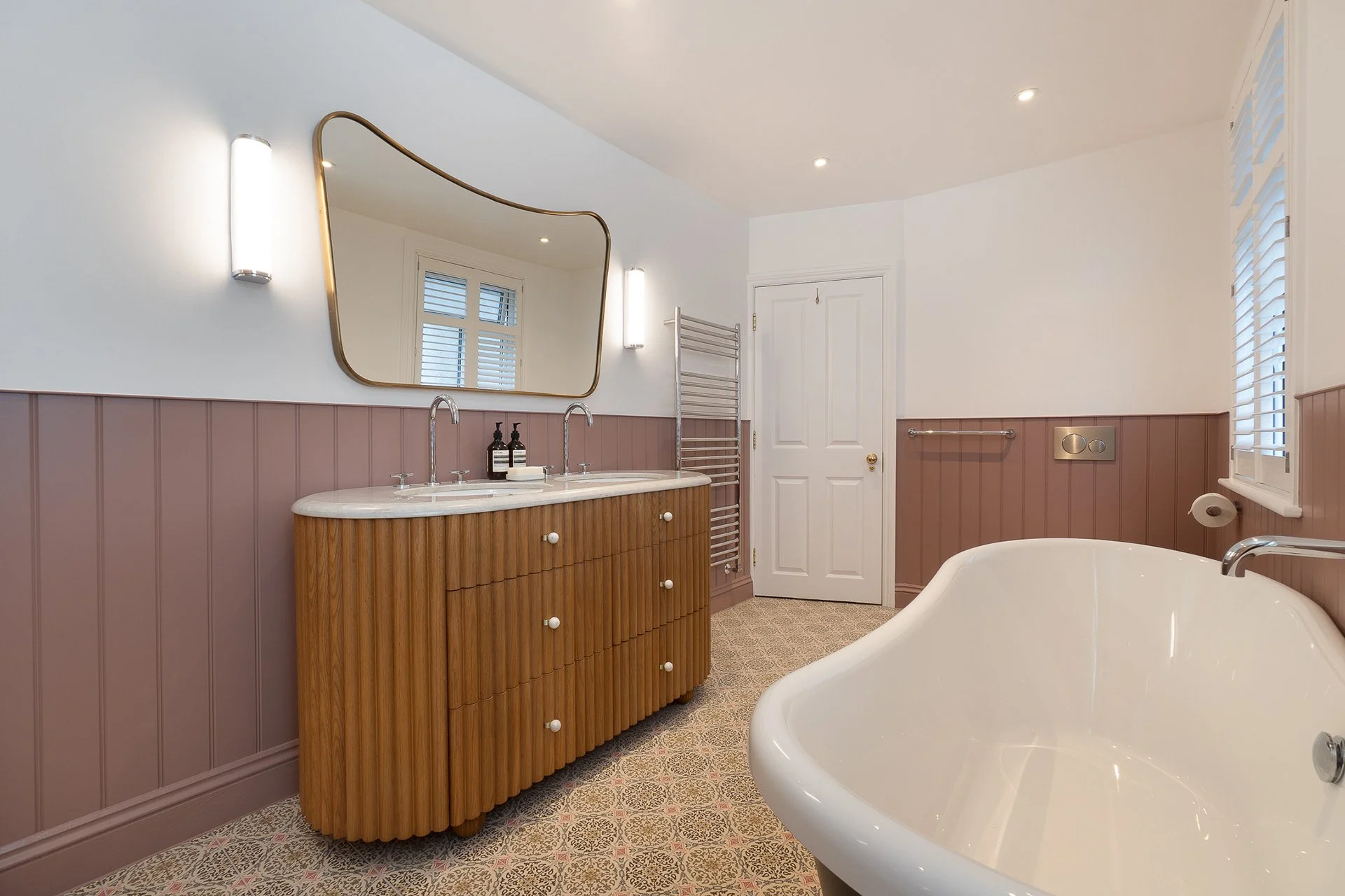 Bathroom with a bathtub, wooden vanity with two sinks, large mirror, wall-mounted towel warmer, toilet paper holder, and window with white shutters.