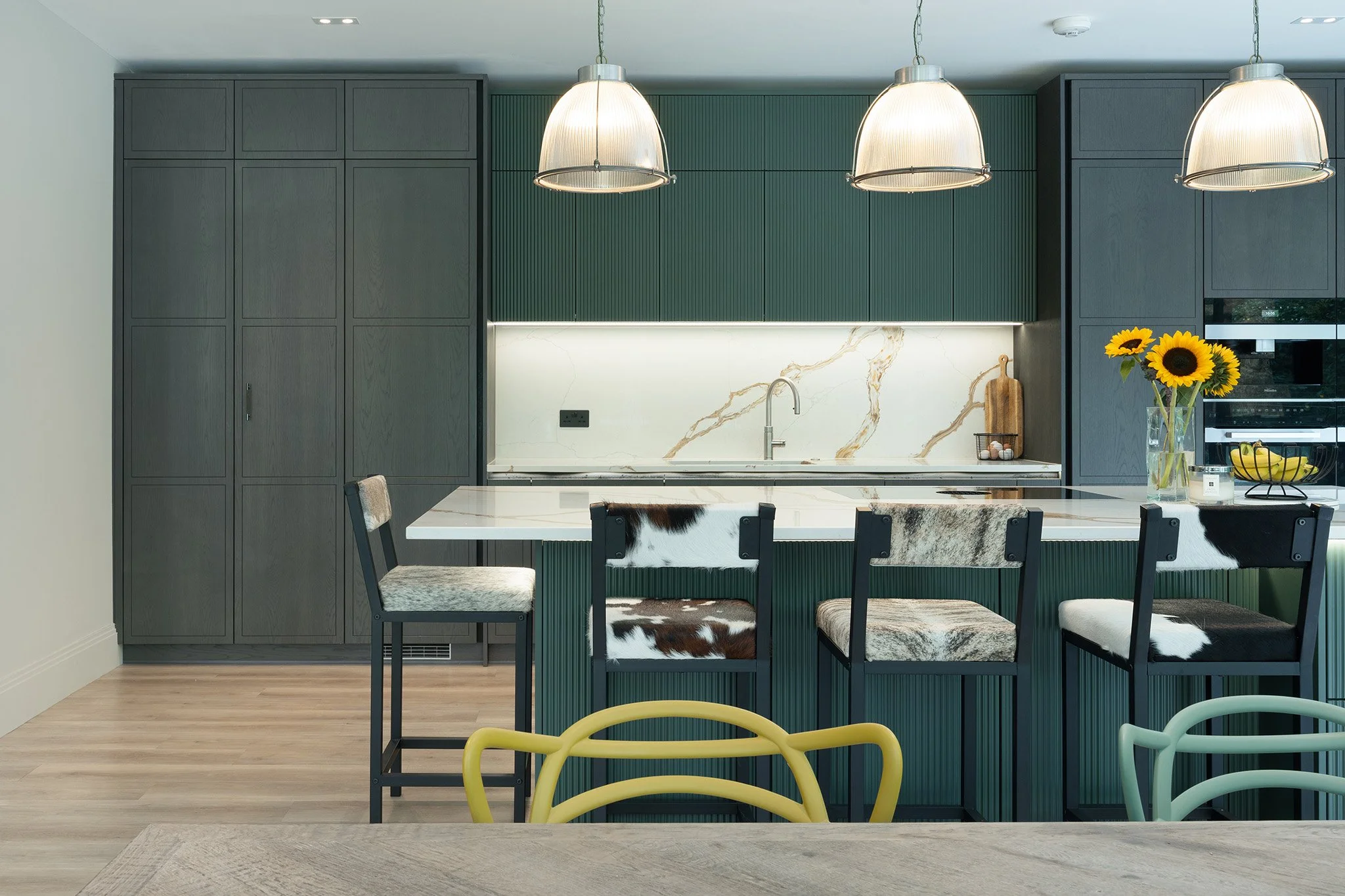 Modern kitchen with dark gray cabinets, white marble countertop, green textured cabinetry, beige bar stools with cowhide upholstery around a white marble island, pendant lights, sunflower vase on the counter, and wooden cutting board.