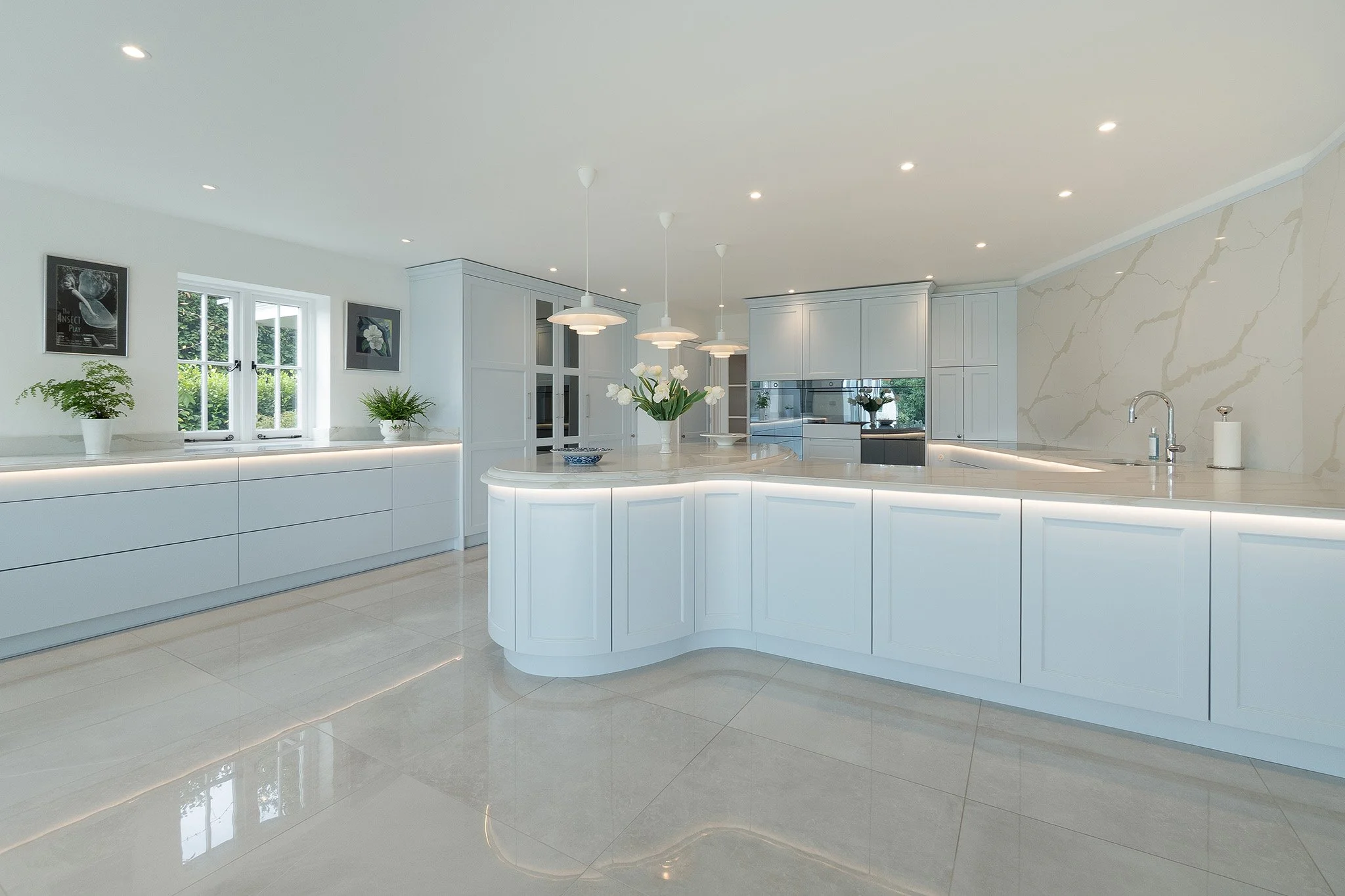 A modern, white kitchen with marble backsplash, pendant lights, and potted plants, featuring a large island and sleek cabinetry.