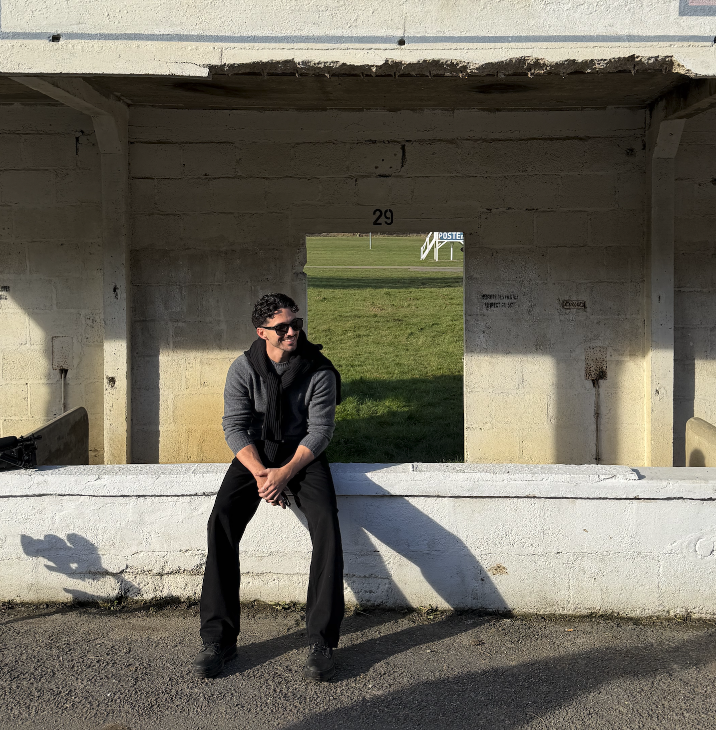 A man sitting on a white wall of a concrete structure with an opening behind him showing green grass and a sign with the number 29.