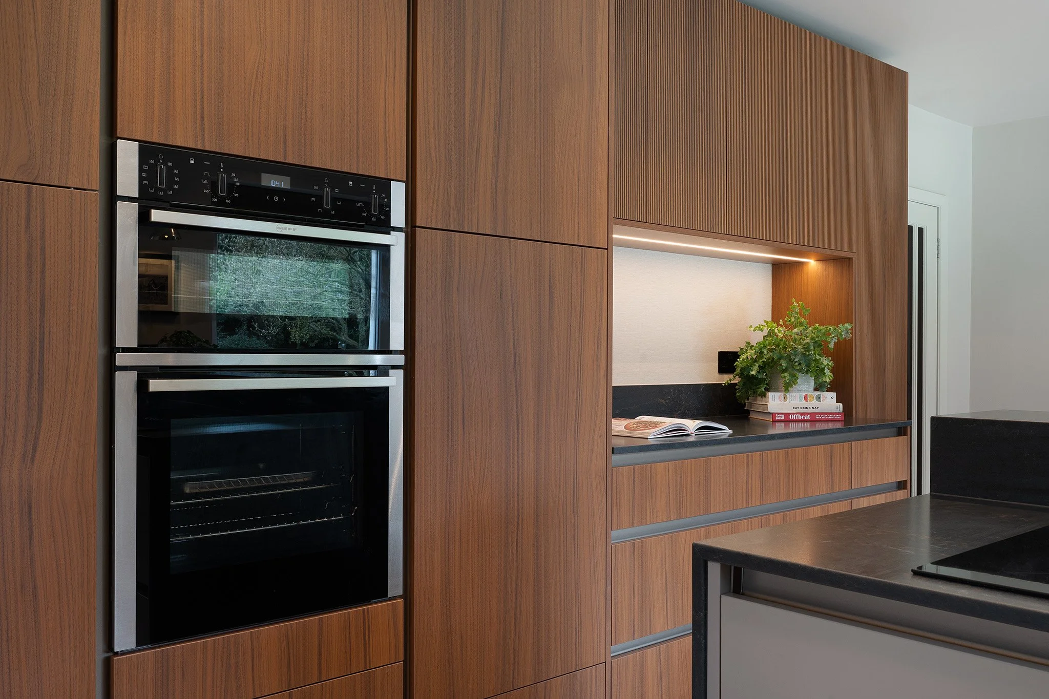 Modern kitchen with wooden cabinets, built-in oven, open shelves with books and a potted plant, and a black countertop.
