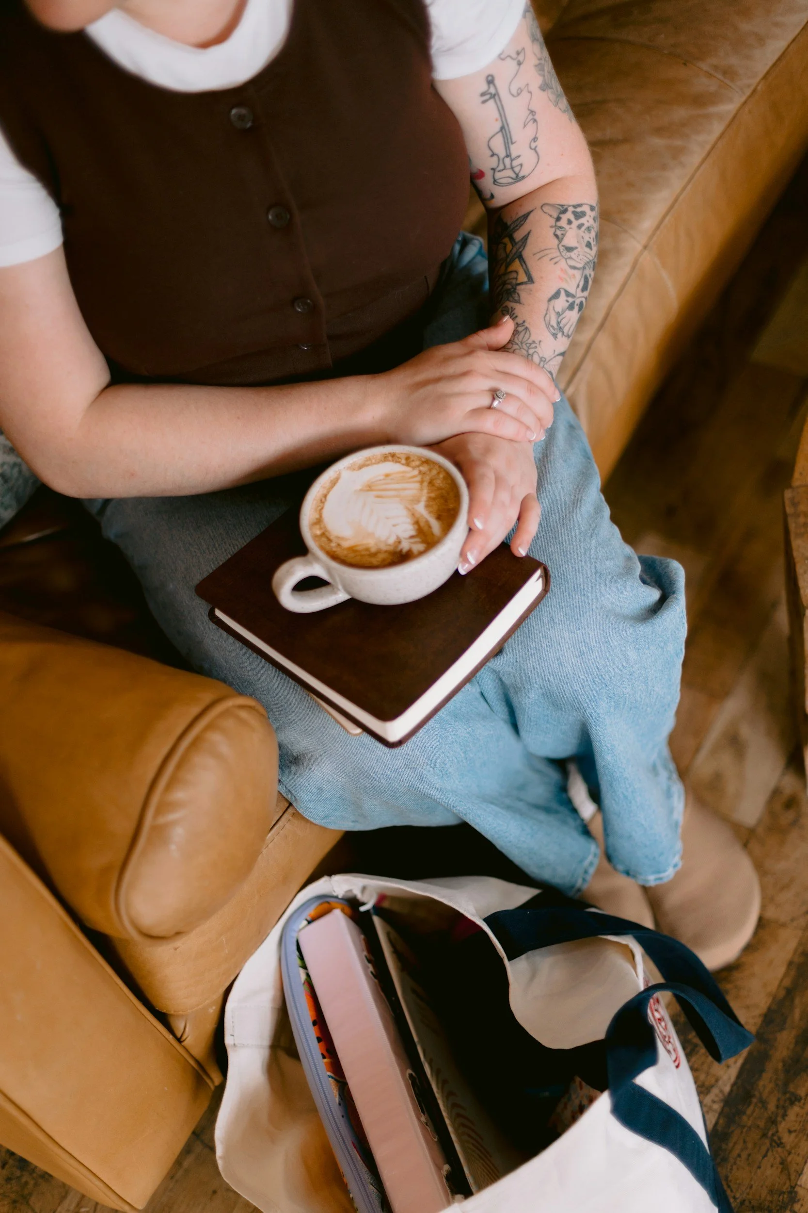 A person sitting on a leather chair with tattooed arms, holding a cup of coffee with latte art, a notebook on their lap, and a bag on the floor beside them.