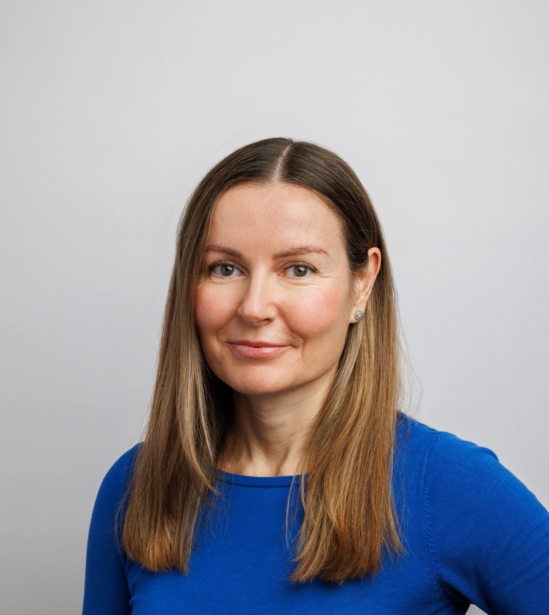 Portrait of a woman with long, light brown hair wearing a blue top, smiling slightly against a plain light gray background.