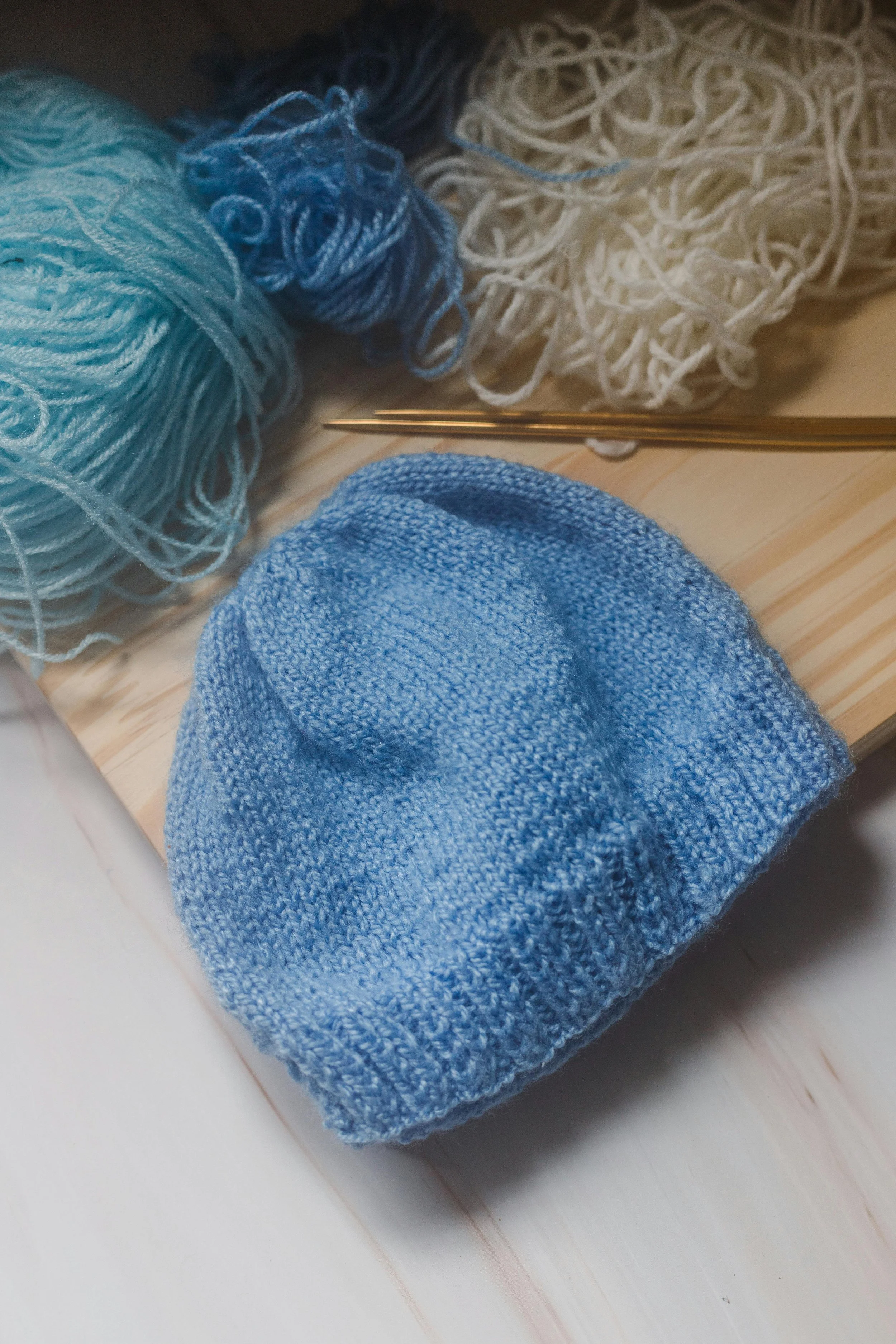 A light blue knitted sock, three balls of yarn in shades of blue and white, and a pair of knitting needles on a wooden surface.