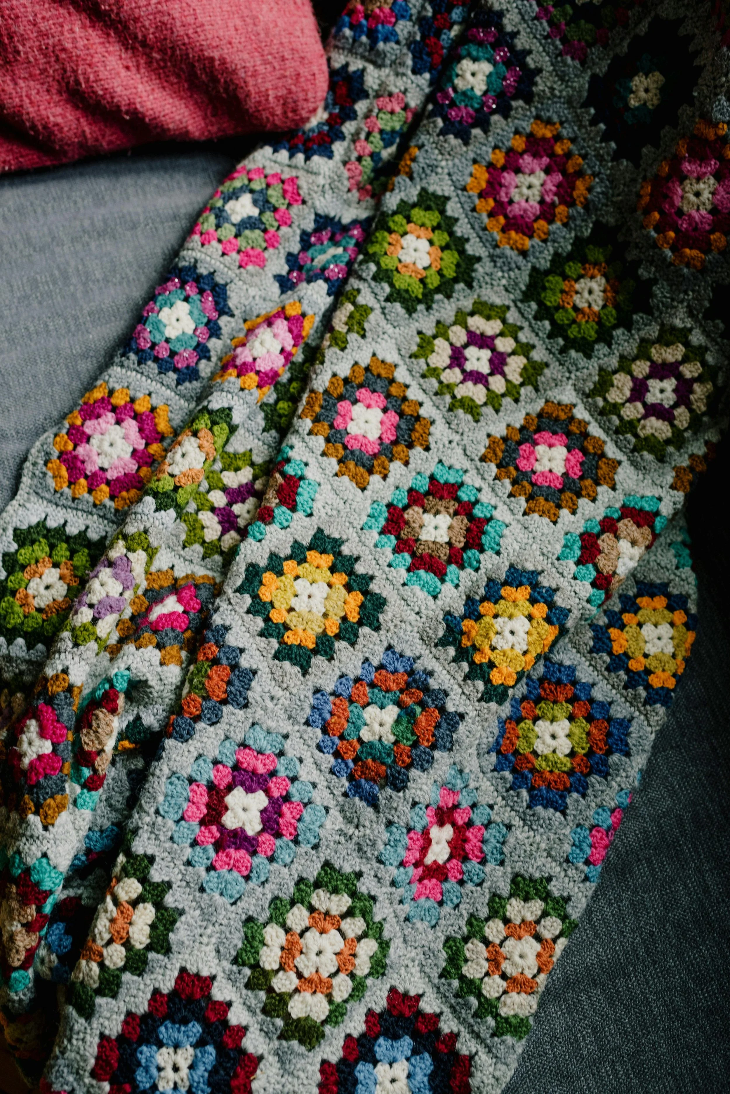 A colorful crochet blanket with multiple floral patterns laid on a surface, with a pink fabric in the upper left corner.