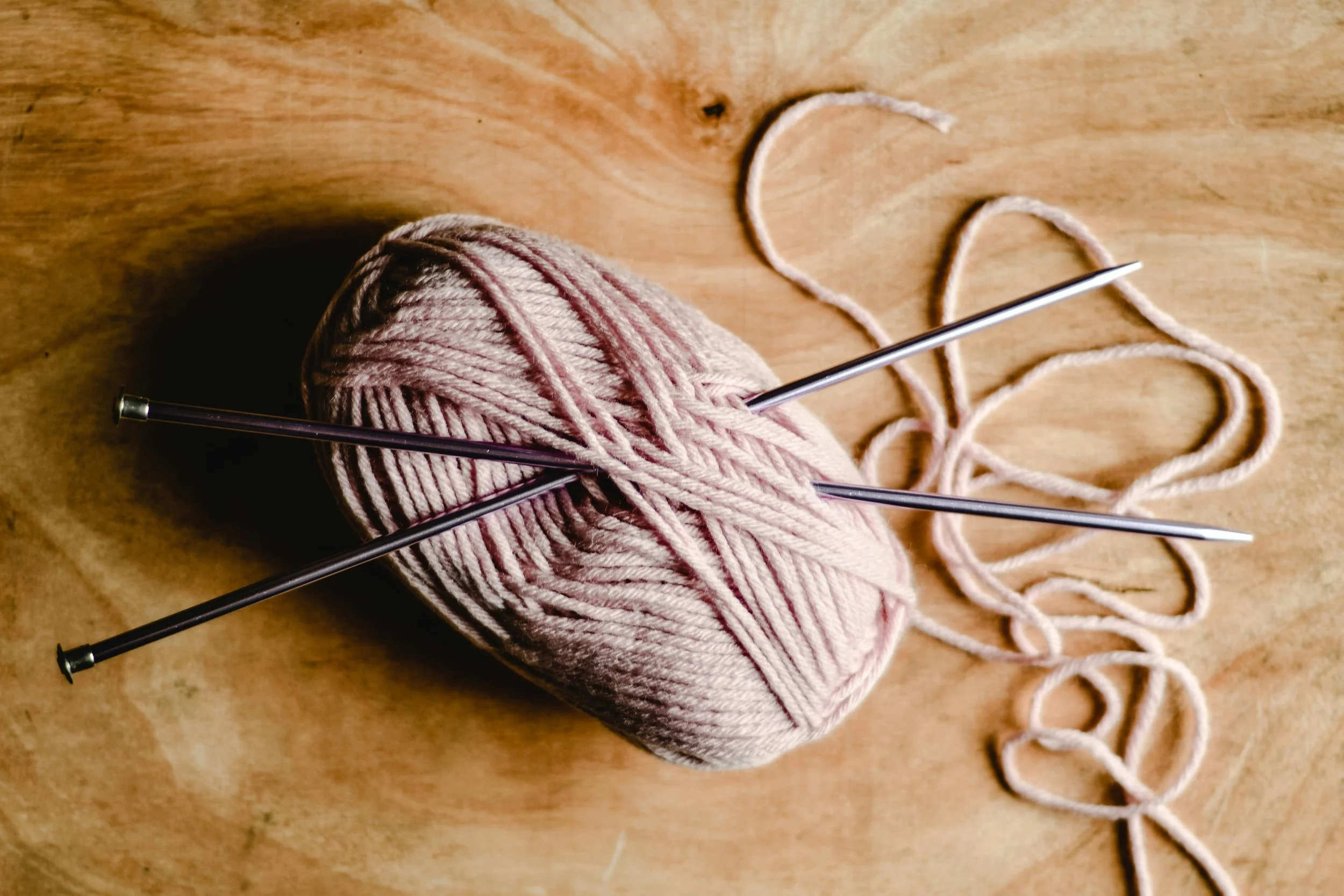 Ball of pink yarn with knitting needles and loose yarn strands on a wooden surface.