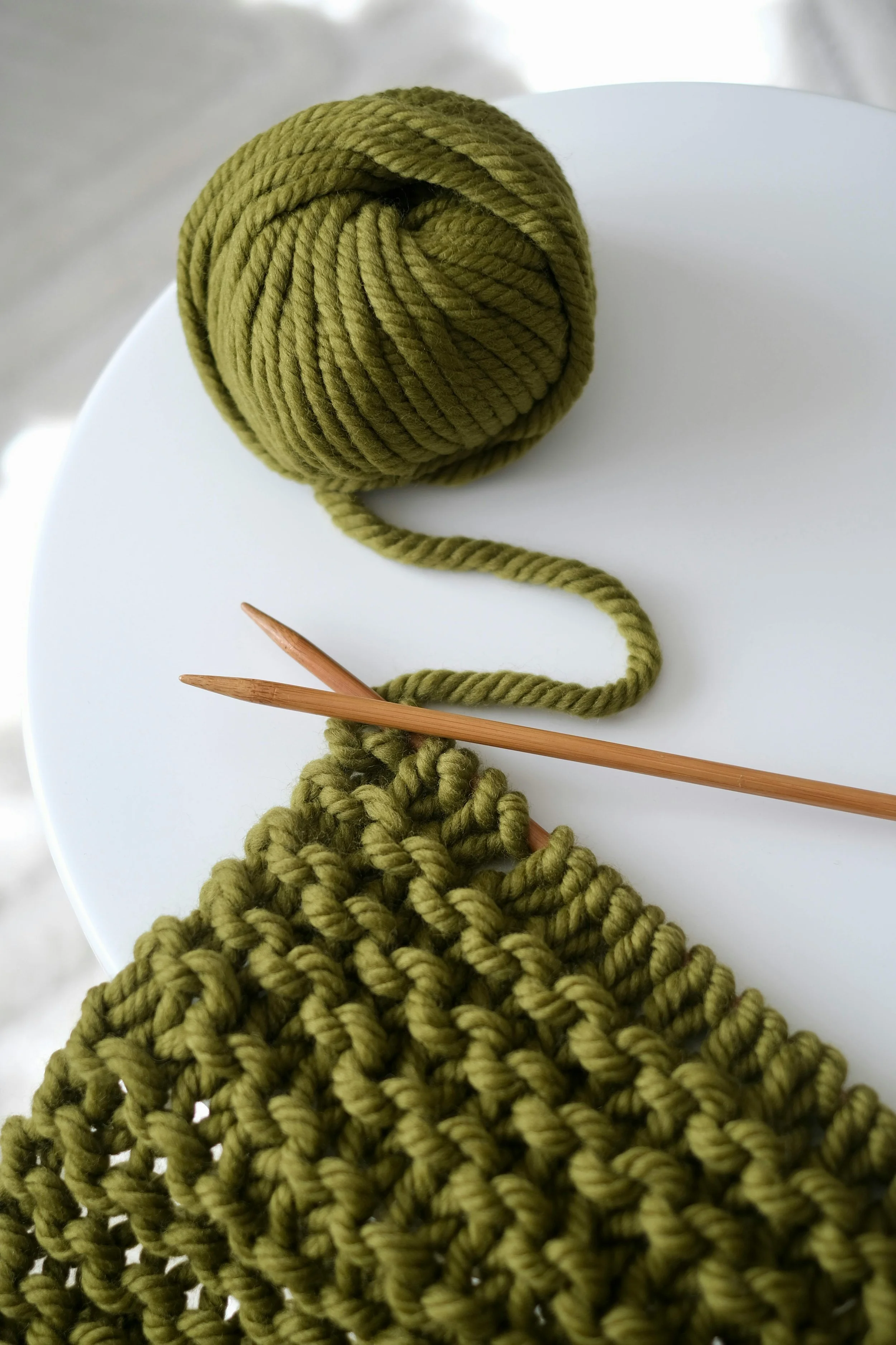 A ball of green yarn, a beige wooden knitting needle, and a partially knitted green fabric are on a white surface.
