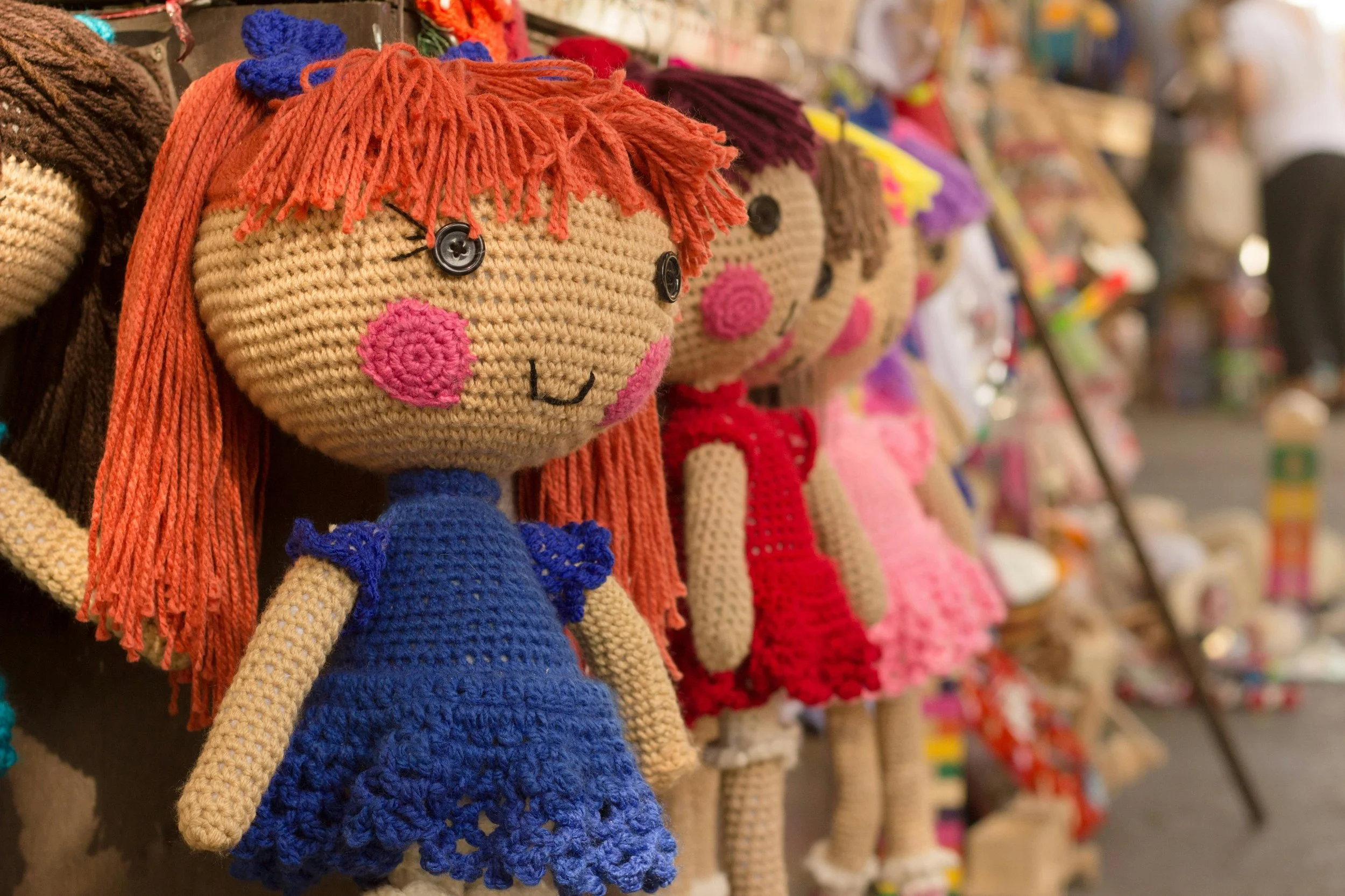 Colorful crochet dolls with button eyes, pink cheeks, and yarn hair on a display shelf at a market.