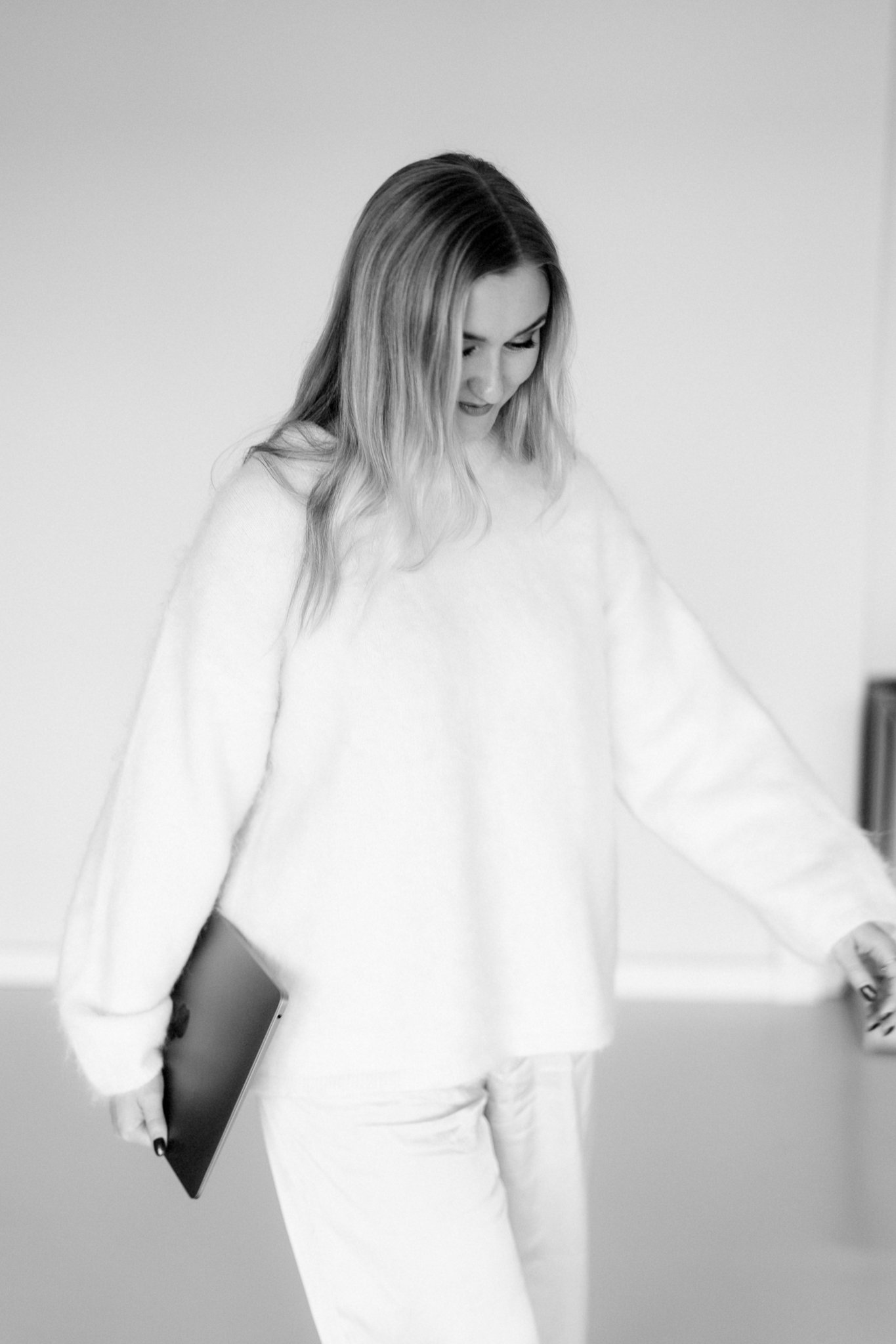 Nainen pukeutuneena valkoiseen, katsoo alas, kädessään puhelin, seisoo harmaalla taustalla.