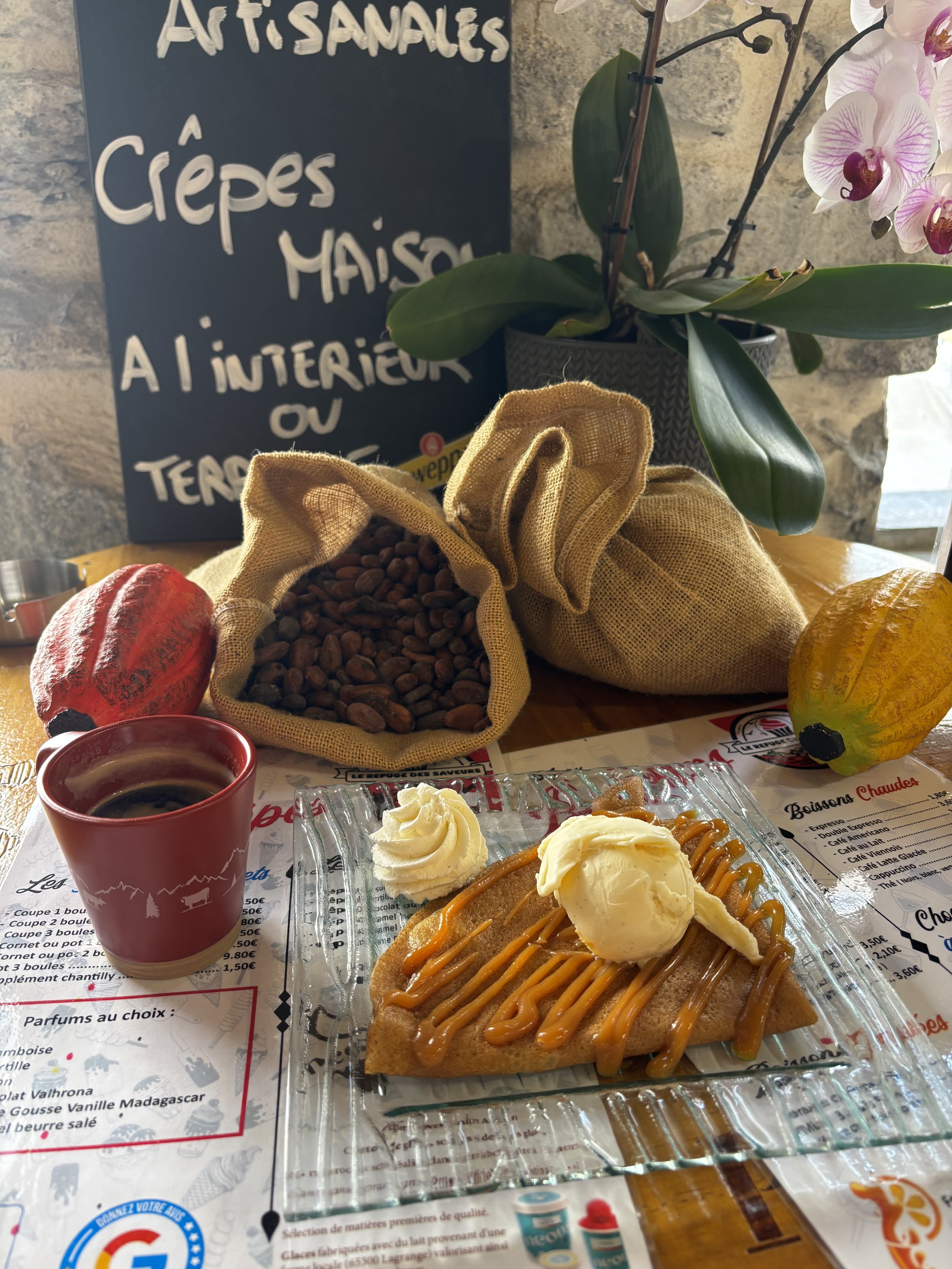 Une assiette de crêpe garnie de caramel, accompagnée de deux boules de glace à la vanille, sur une table dans un restaurant. Une tasse de café, un sac de cacao, et une citrouille en décoration sont visibles, avec un panneau indiquant des artisanaux, crêpes maison, à l'intérieur ou en terrasse, et un orchidée en arrière-plan.