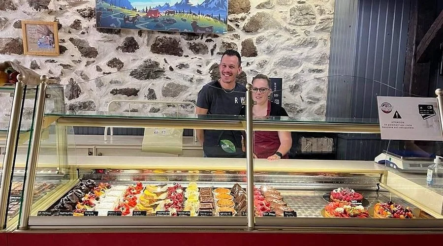 Deux personnes derrière un comptoir de pâtisserie dans une boutique, avec des gâteaux et desserts exposés dans une vitrine.