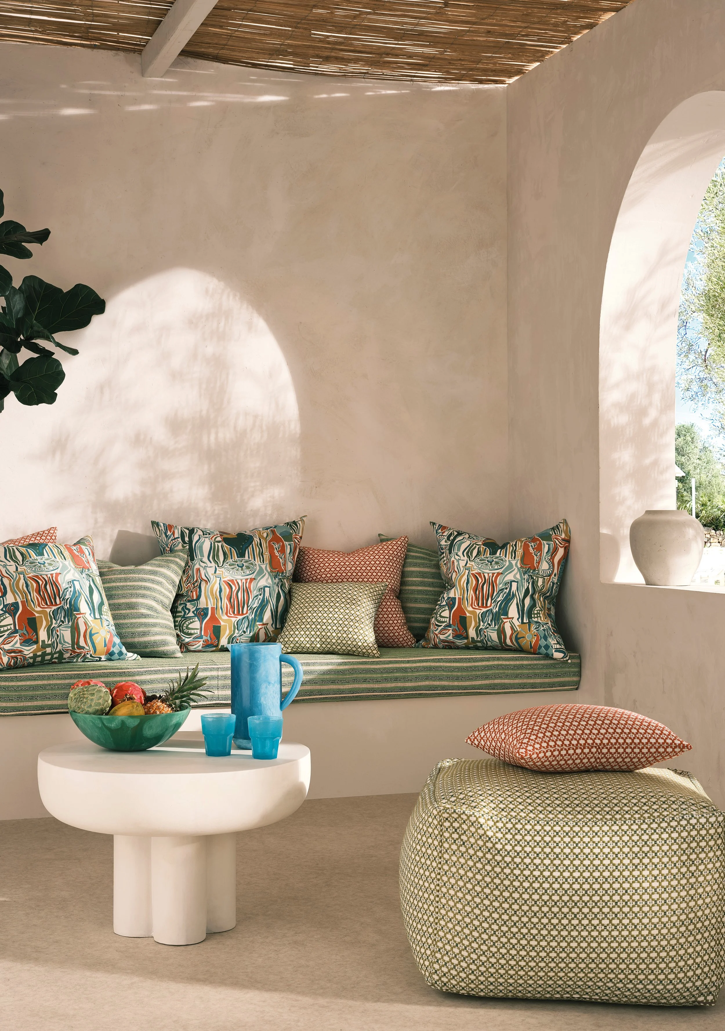 Brightly lit interior with white plaster walls, a built-in cushioned bench with colorful patterned pillows, a small round white side table with a bowl of fruit and blue ceramics, and a large patterned pouf with a pillow, with a large white vase on th