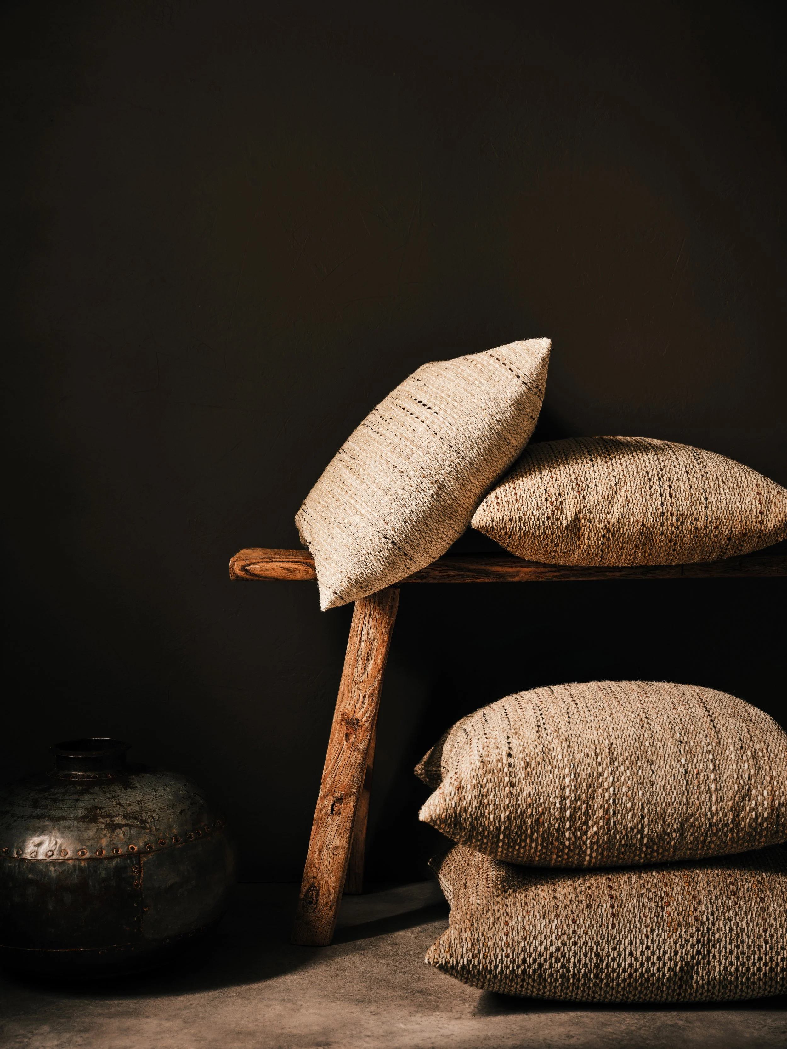 Stack of three textured beige cushions on a wooden bench against a black wall, with a dark metallic decorative vase on the floor nearby.