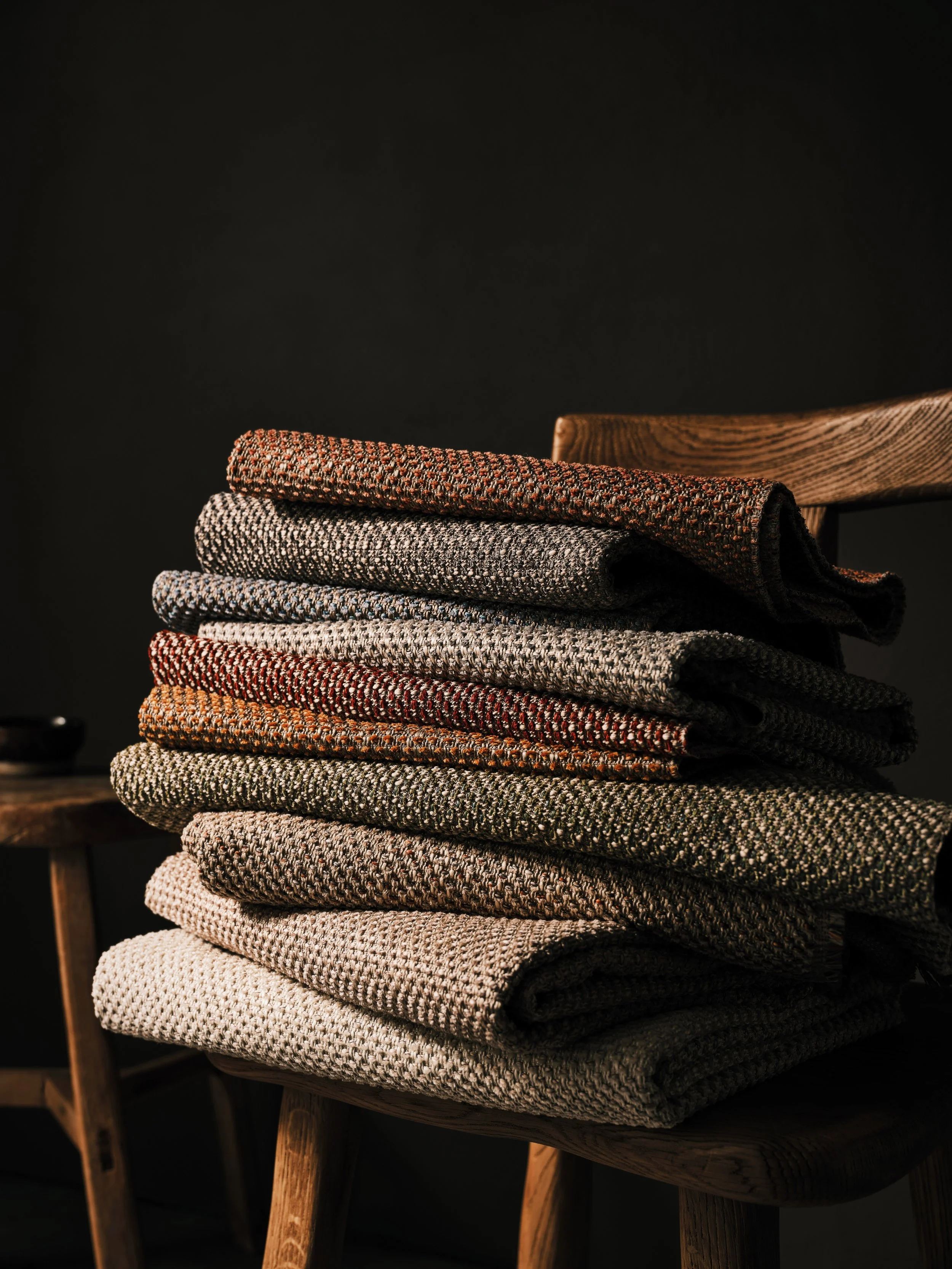 A stack of folded, textured, multicolored woolen blankets on a wooden chair against a dark background.