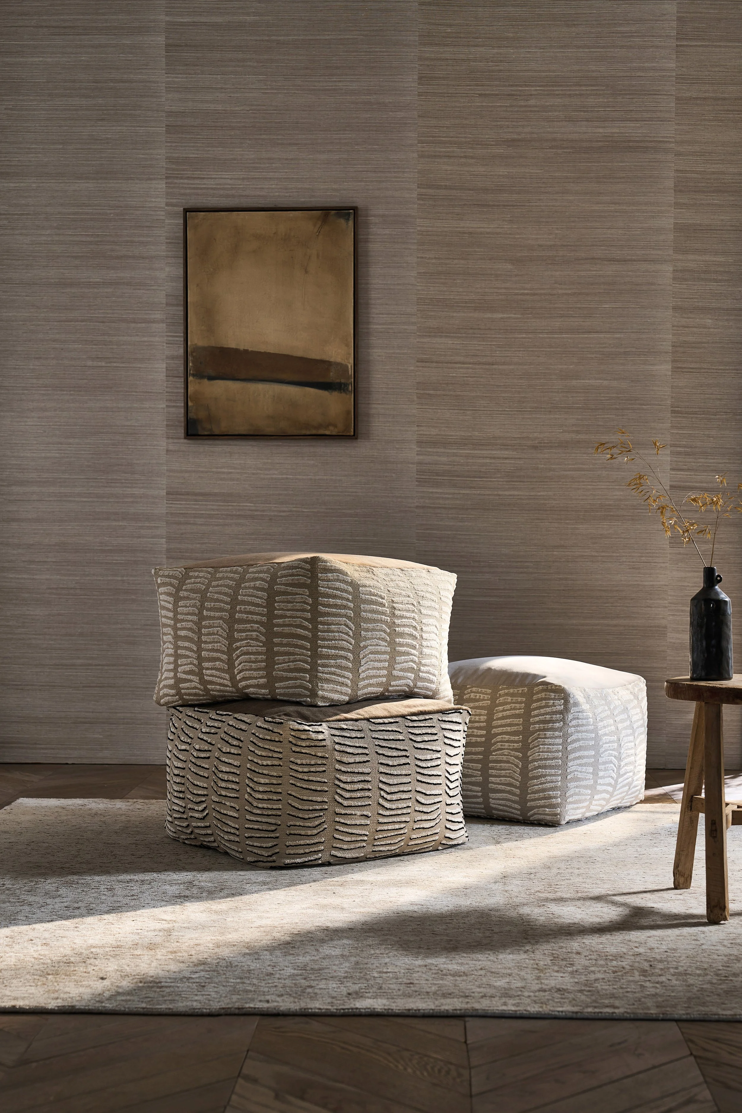 Minimalist living space with two textured fabric poufs, a wooden side table with a black vase and dried branches, a neutral-toned abstract painting, and a beige rug on dark hardwood floor.