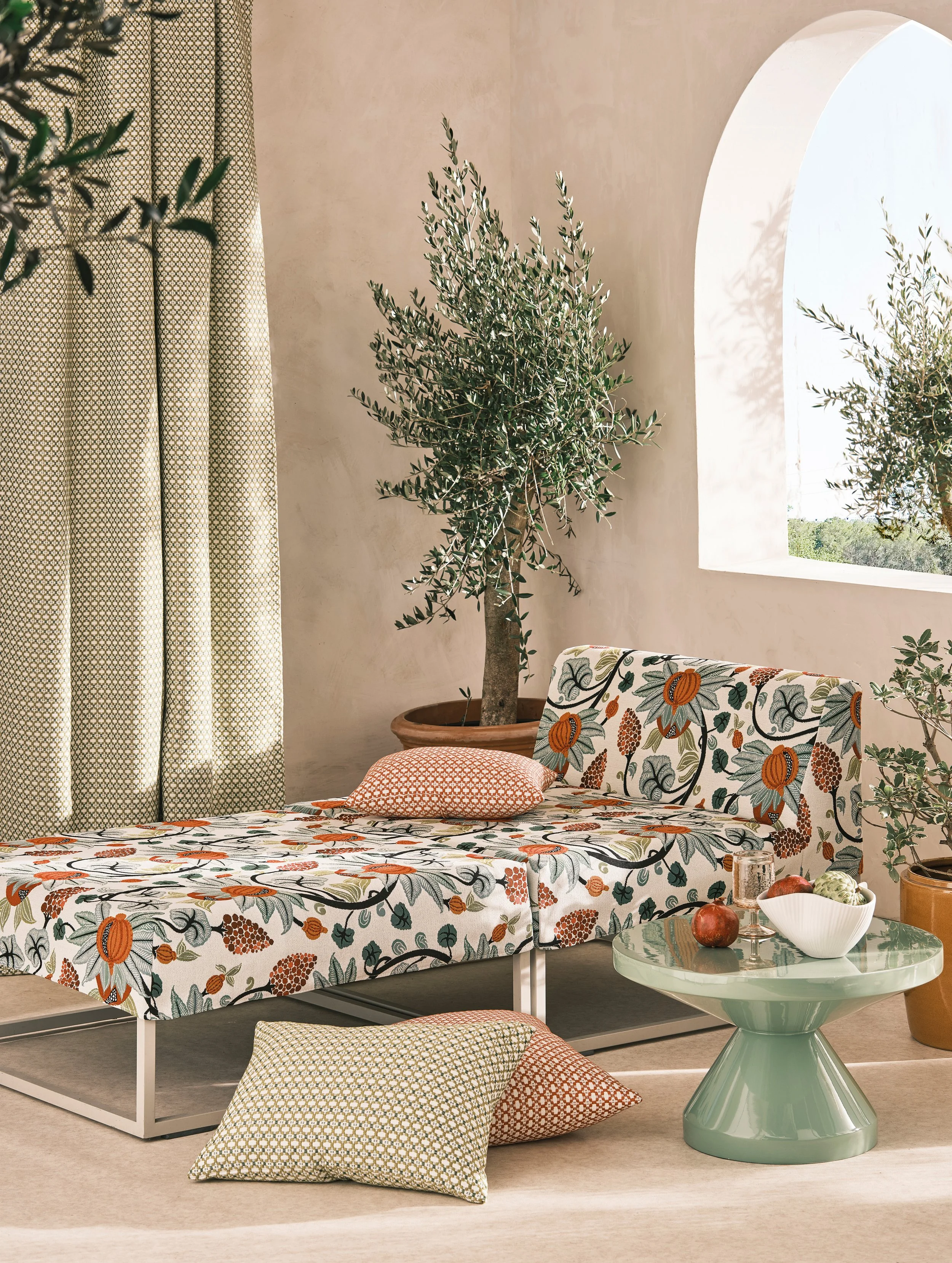 A living room scene with a floral-patterned daybed, a green side table with decorative items, potted plants, and a window letting in natural light, with beige walls, a curtain, and a beige carpet.