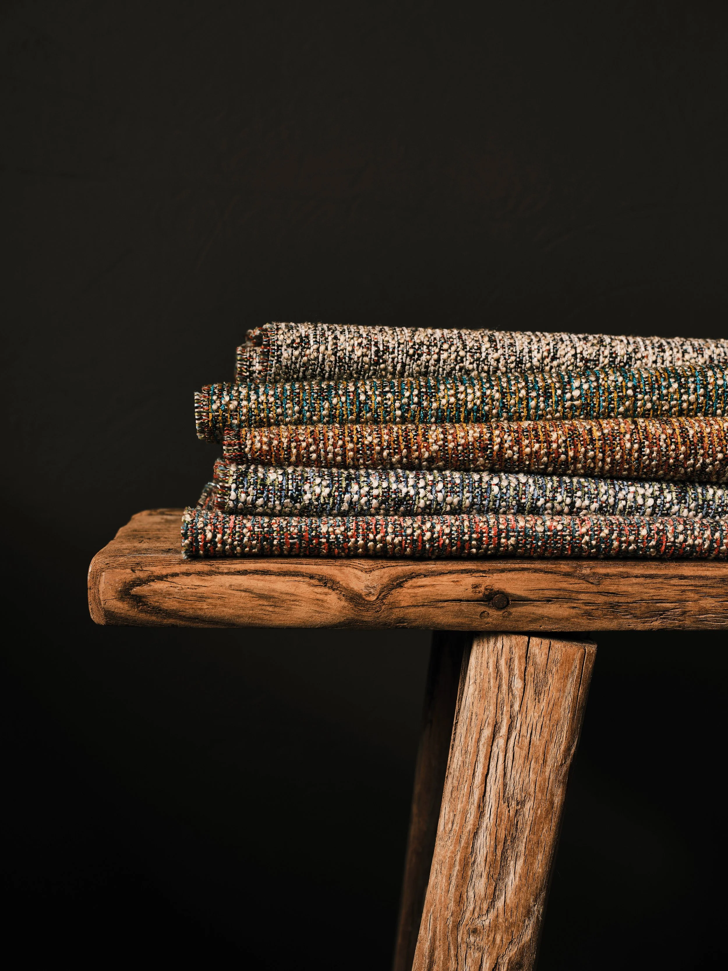 Stack of patterned textiles on a wooden stool against a black background.