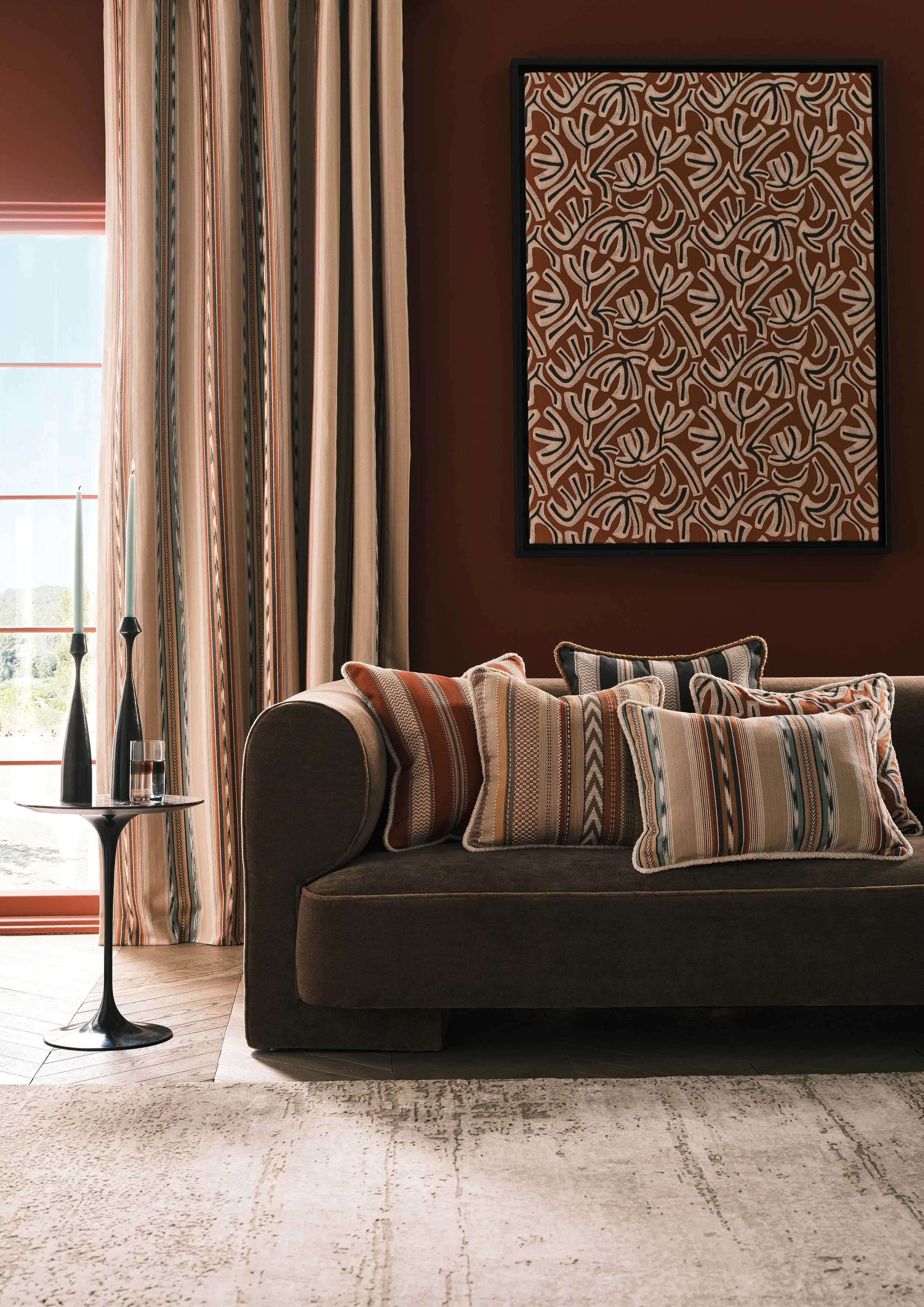 Living room with a dark brown sofa, striped throw pillows, a side table with candles and a glass of water, a large window with beige and orange curtains, and an abstract patterned artwork on a dark red wall.