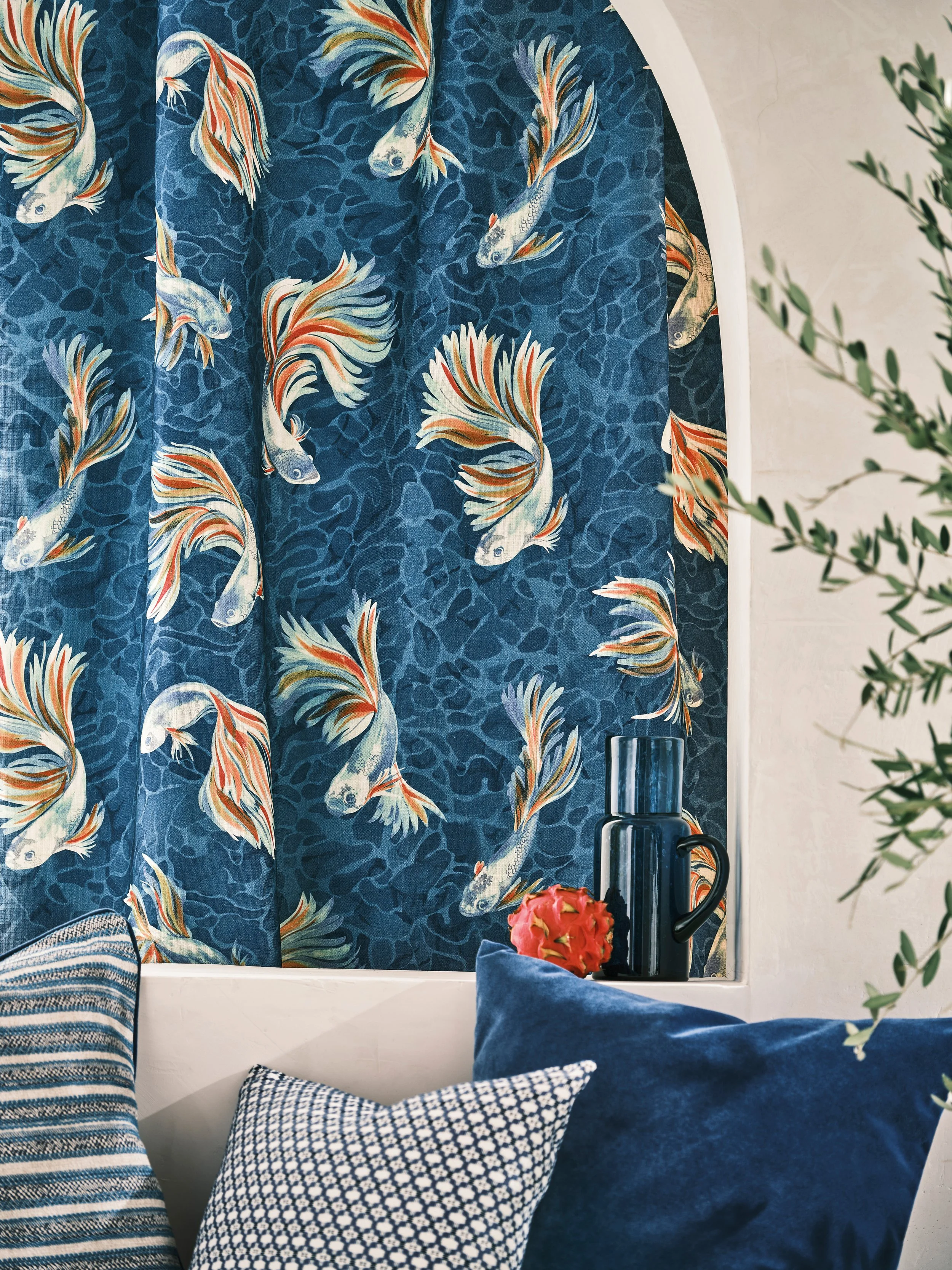 A window with a blue curtain decorated with fish pattern, a ledge with a black pitcher, a pink decorative fruit, and two pillows in front of the window, one striped and one navy blue.
