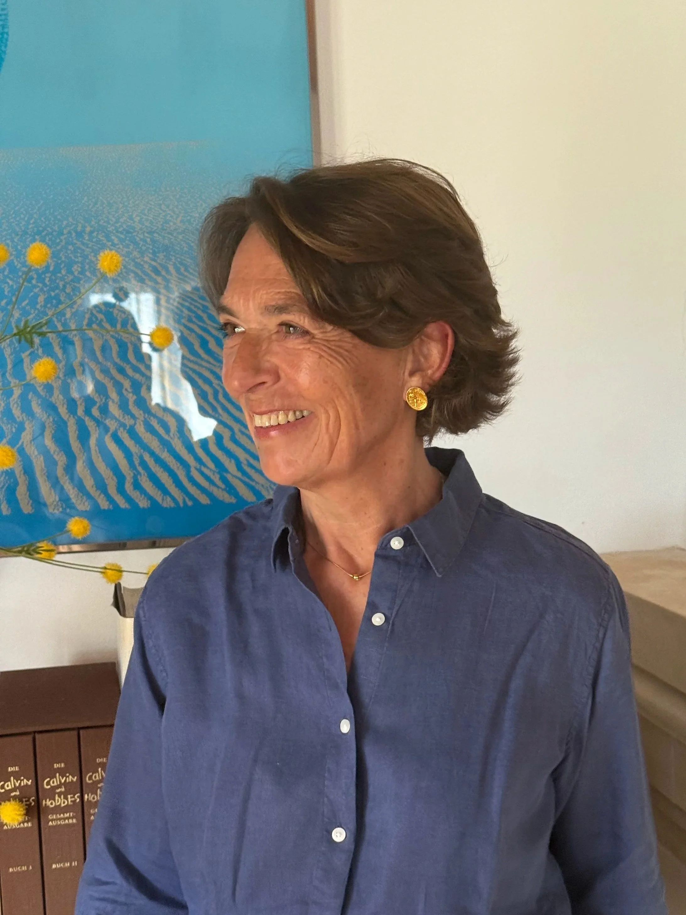 A woman with short brown hair smiling, wearing a blue button-up shirt and gold earrings, standing indoors with a colorful abstract painting and a stack of books in the background.