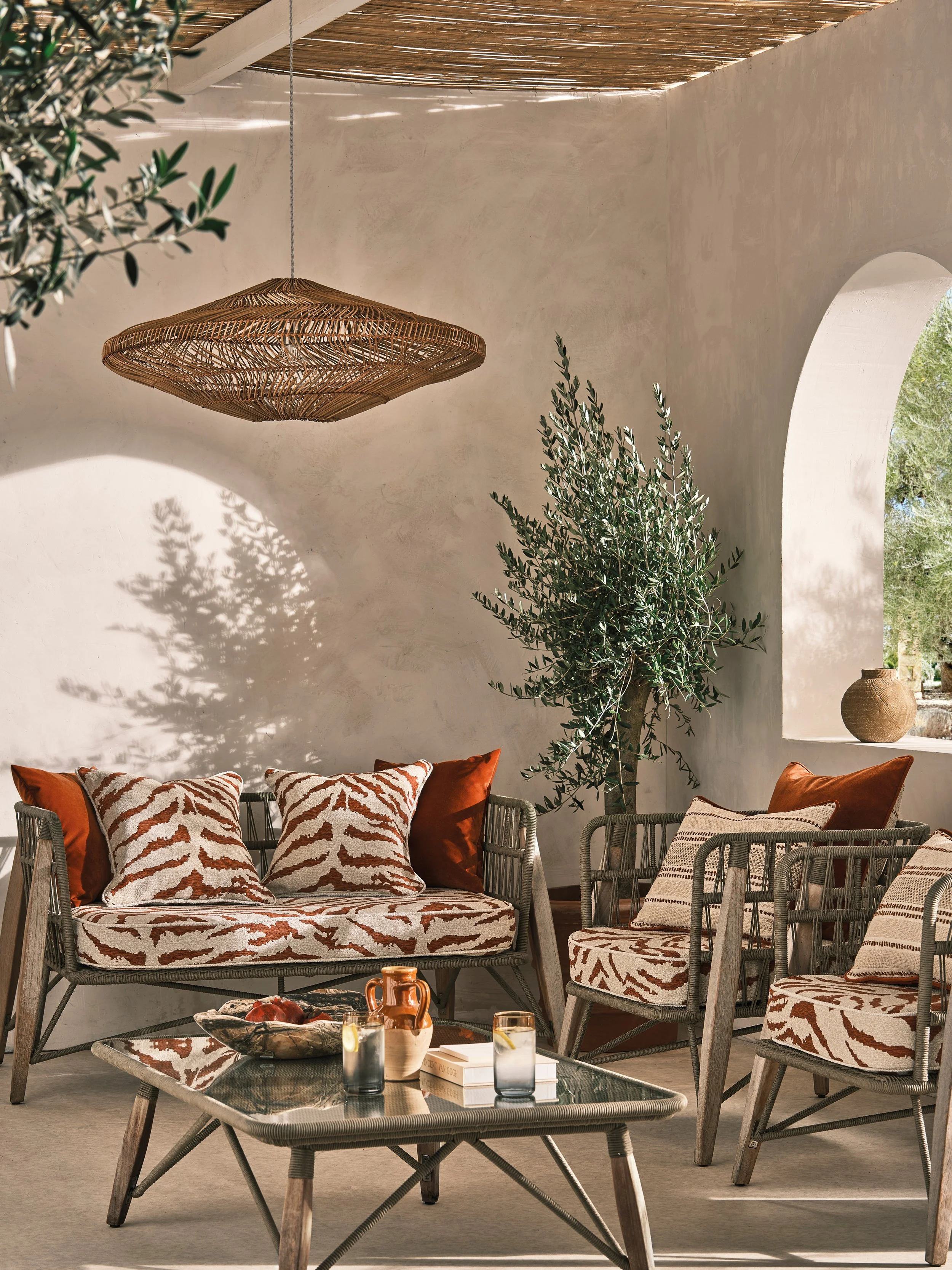 A cozy outdoor patio with a wicker sofa and chairs featuring animal print cushions, a glass-topped coffee table with drinks and snacks, a large potted tree, and a woven ceiling light, all illuminated by natural sunlight.