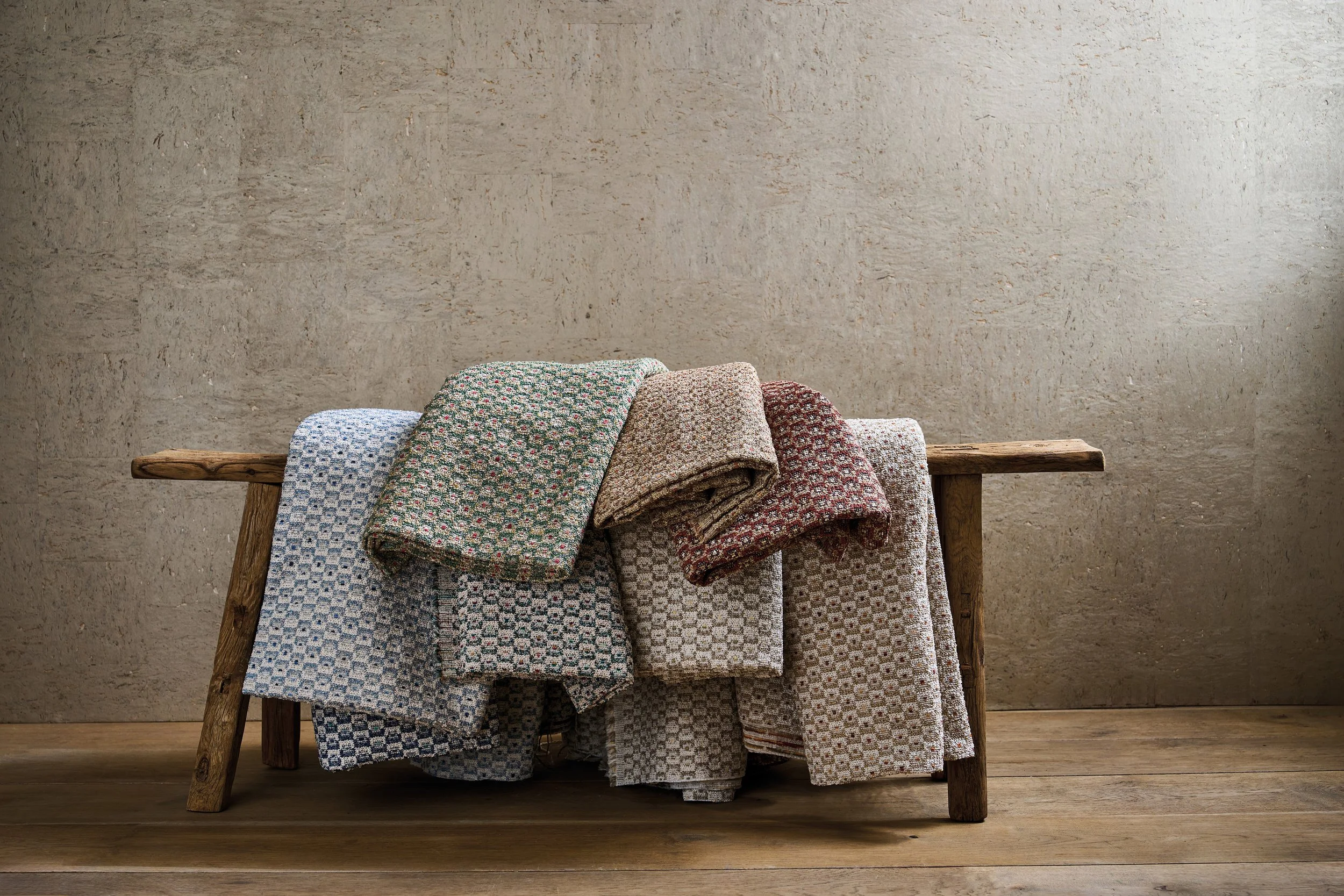 A wooden bench with several folded and draped textured blankets in various patterned fabric on top, set against a plain beige wall and on a wooden floor.