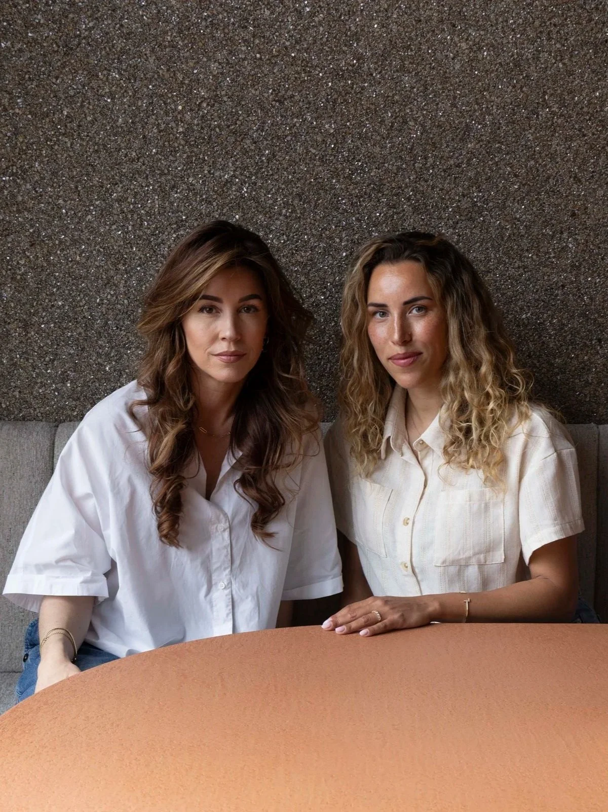 Twee vrouwen zitten naast elkaar aan een tafel, gekleed in lichte shirts, met een sparkelende donkere achtergrond en een beige tafel voor zich.