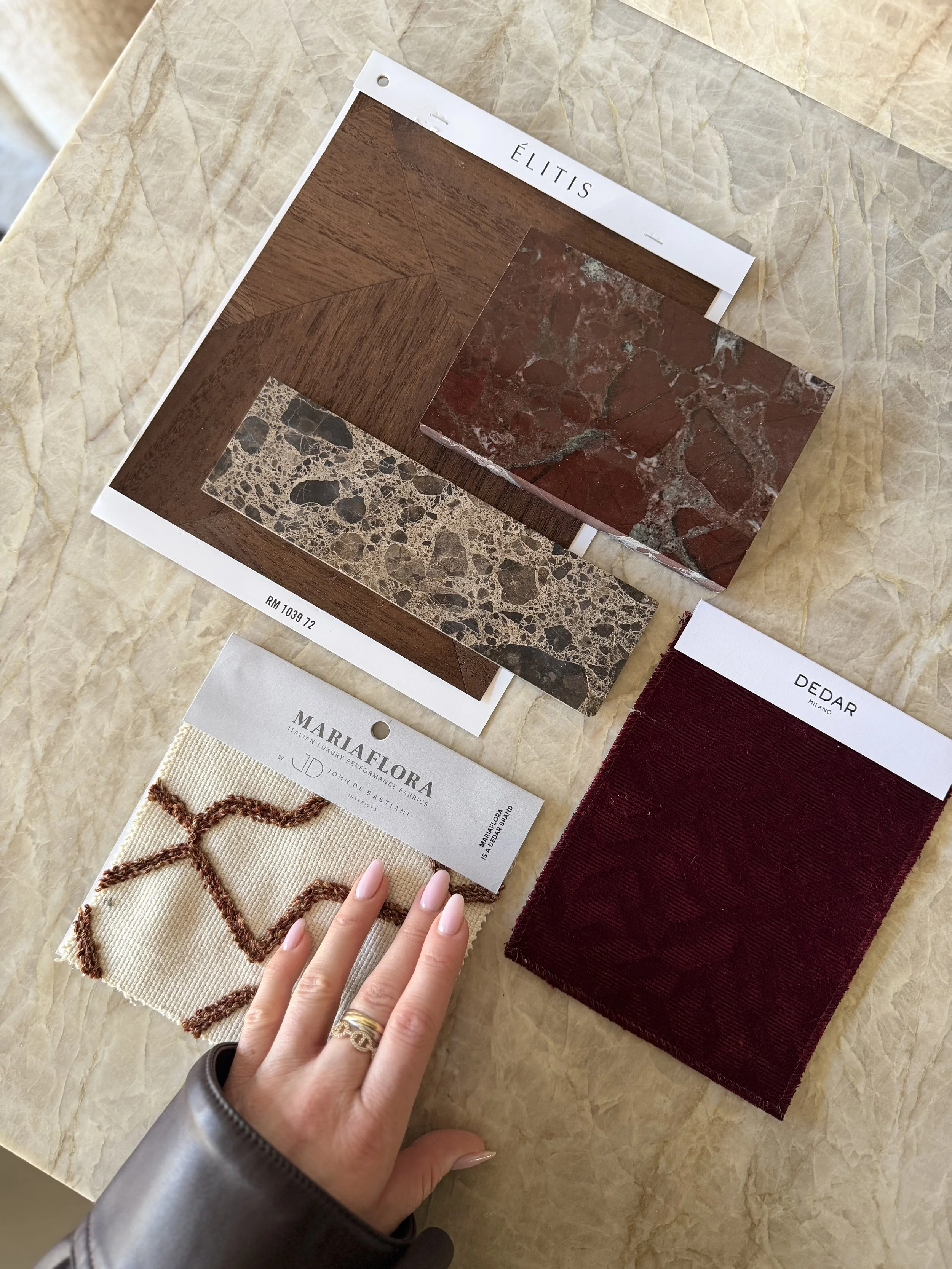 Diverse materialen en kleuren van vloer- en wandbekleding, waaronder keramische tegels, houtprint en stoffen, liggen op een tafel. Een hand met sieraden wijst naar een specifiek monster.