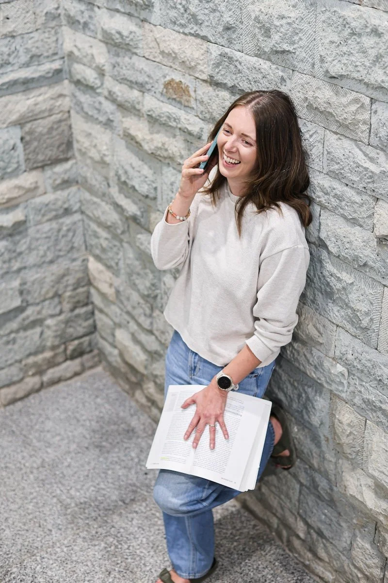 Ellie is leaning against a stone wall, talking on her phone and smiling. She is holding an open book on her right knee and wearing casual clothing.