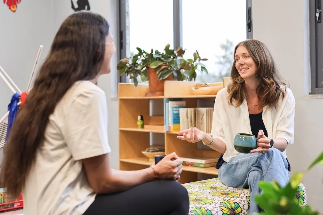 Two women sitting and talking inside a school, Ellie is holding a mug and gesturing, the other woman is listening.