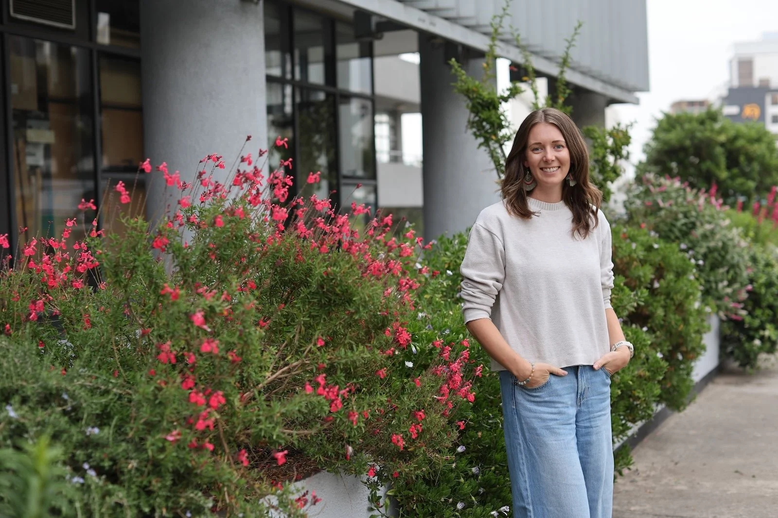 Ellie is wearing a light gray sweatshirt and blue jeans, standing outdoors beside pink and white flowering bushes, smiling at the camera.