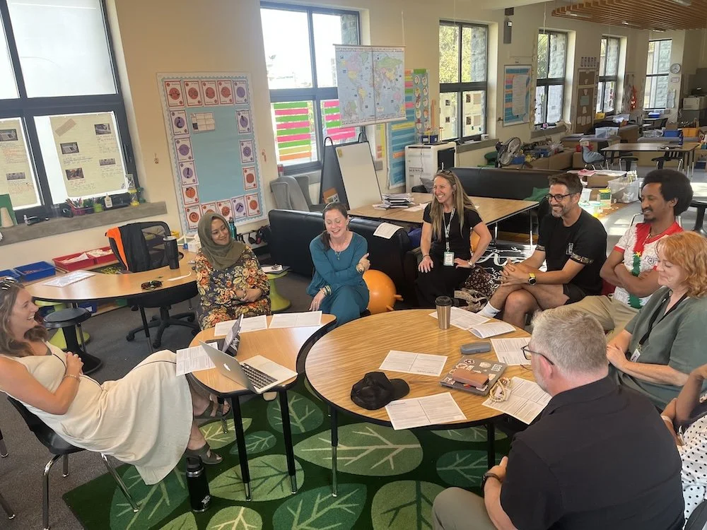 A diverse group of adults engaged in a leadership workshop with Ellie. They are casually sitting around a circular table with laptops, papers, and drinks.