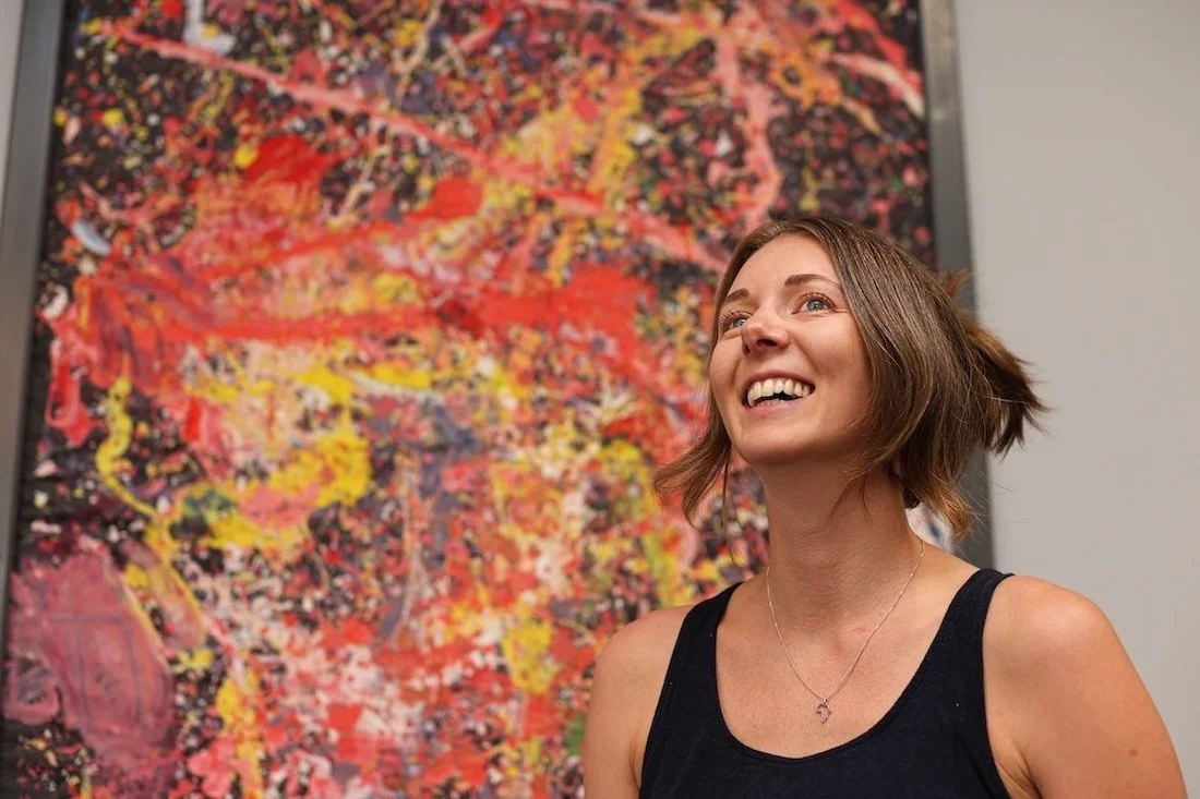 Ellie is smiling and looking up, standing in front of an abstract red and yellow painting.