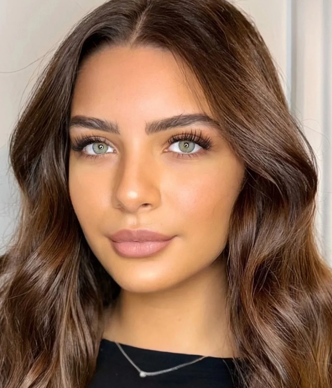 Portrait d'une femme avec des cheveux bruns ondulés, peau claire, maquillage subtil, portant une chaîne fine, fond neutre.