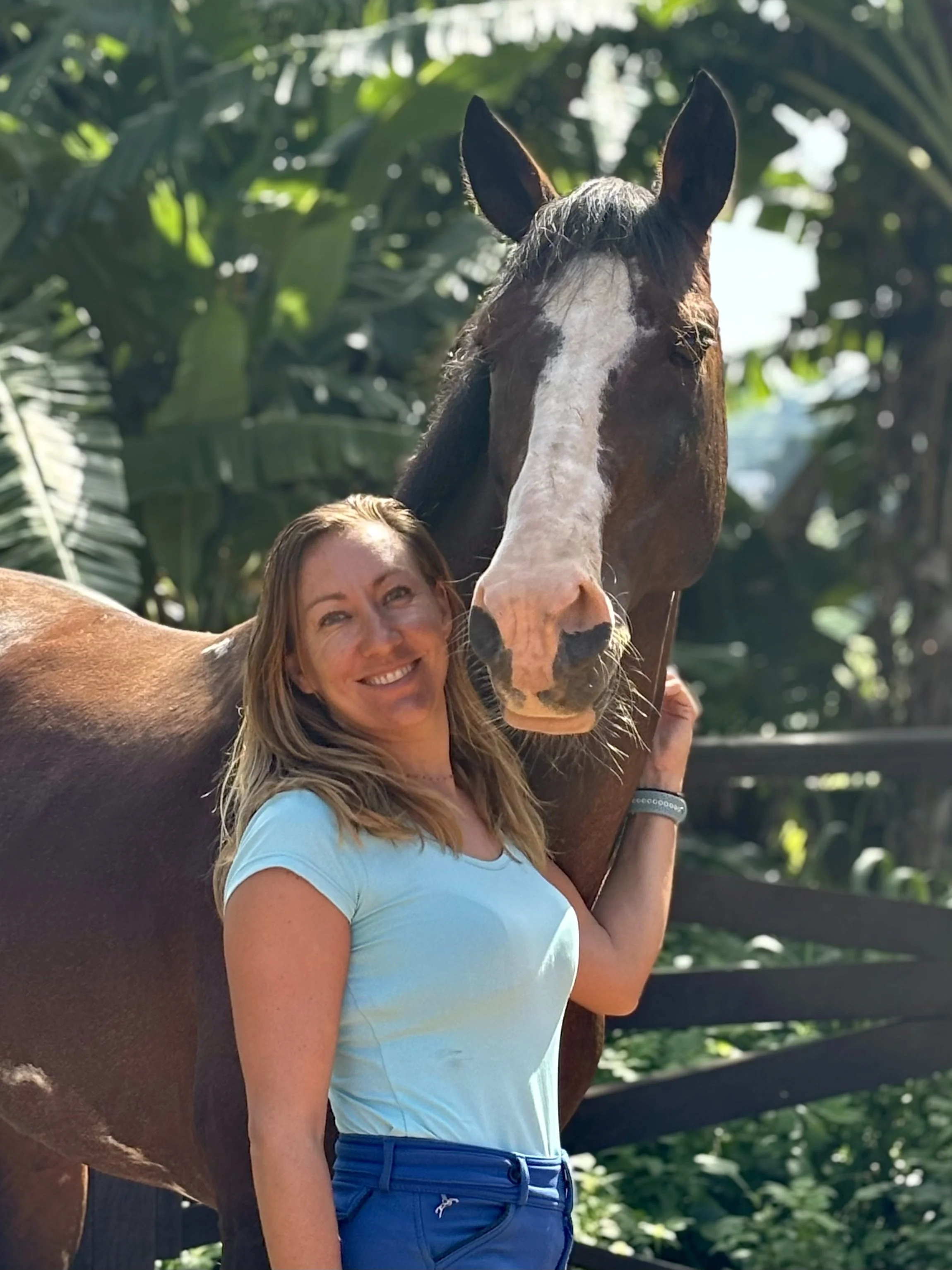 Une femme souriante avec un t-shirt bleu, se tenant près d'un cheval brun avec une tache blanche sur la tête, dans un environnement verdoyant avec des arbres