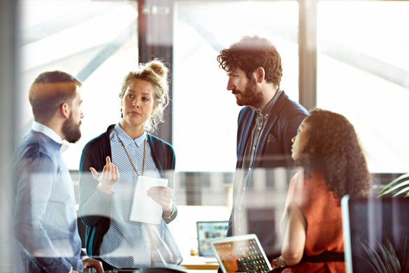 Four diverse professionals engage in a discussion in a modern office with large windows, some holding digital tablets and laptops.