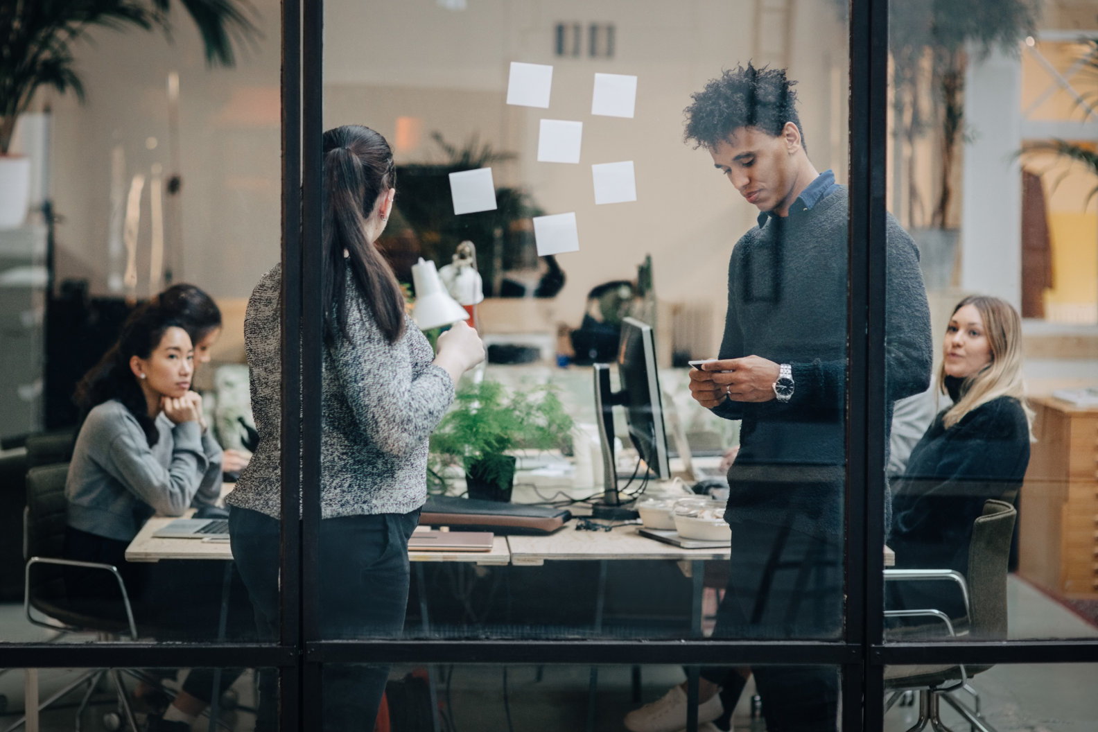 People working at a meeting in a glass-walled office conference room, with sticky notes on the glass and laptops on the table.