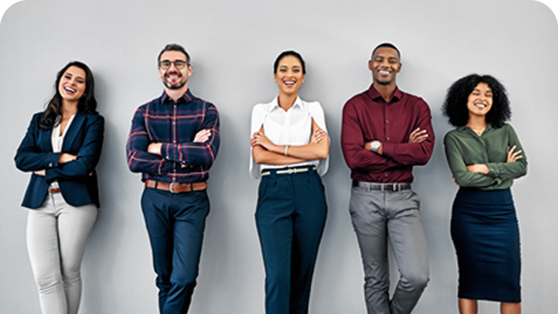 Group of five diverse professionals standing against a plain wall, smiling, with arms crossed.