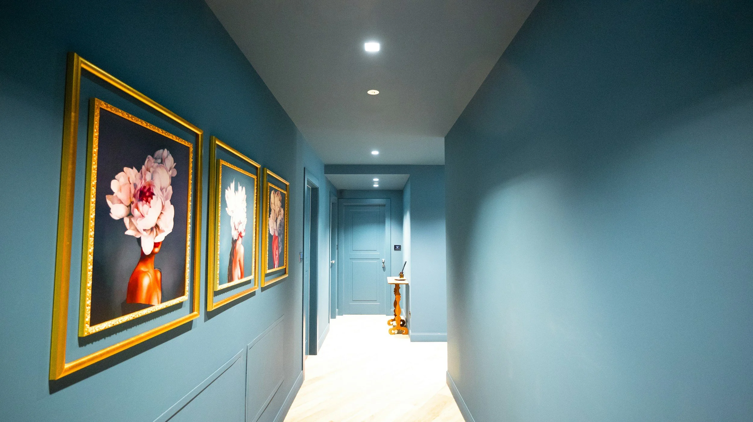 A hallway with blue walls decorated with four framed floral artwork pieces. At the end of the hall, there's a small table with items on top and a door on the right side.