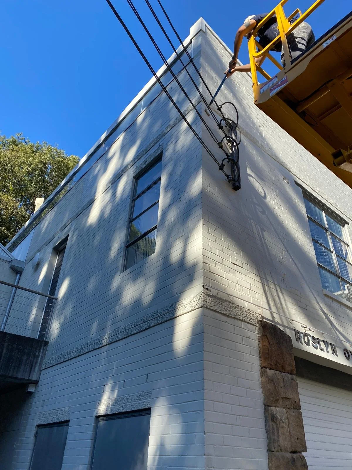 A construction worker on a yellow lift cleaning or repairing power lines on the corner of a white brick building under a clear blue sky.