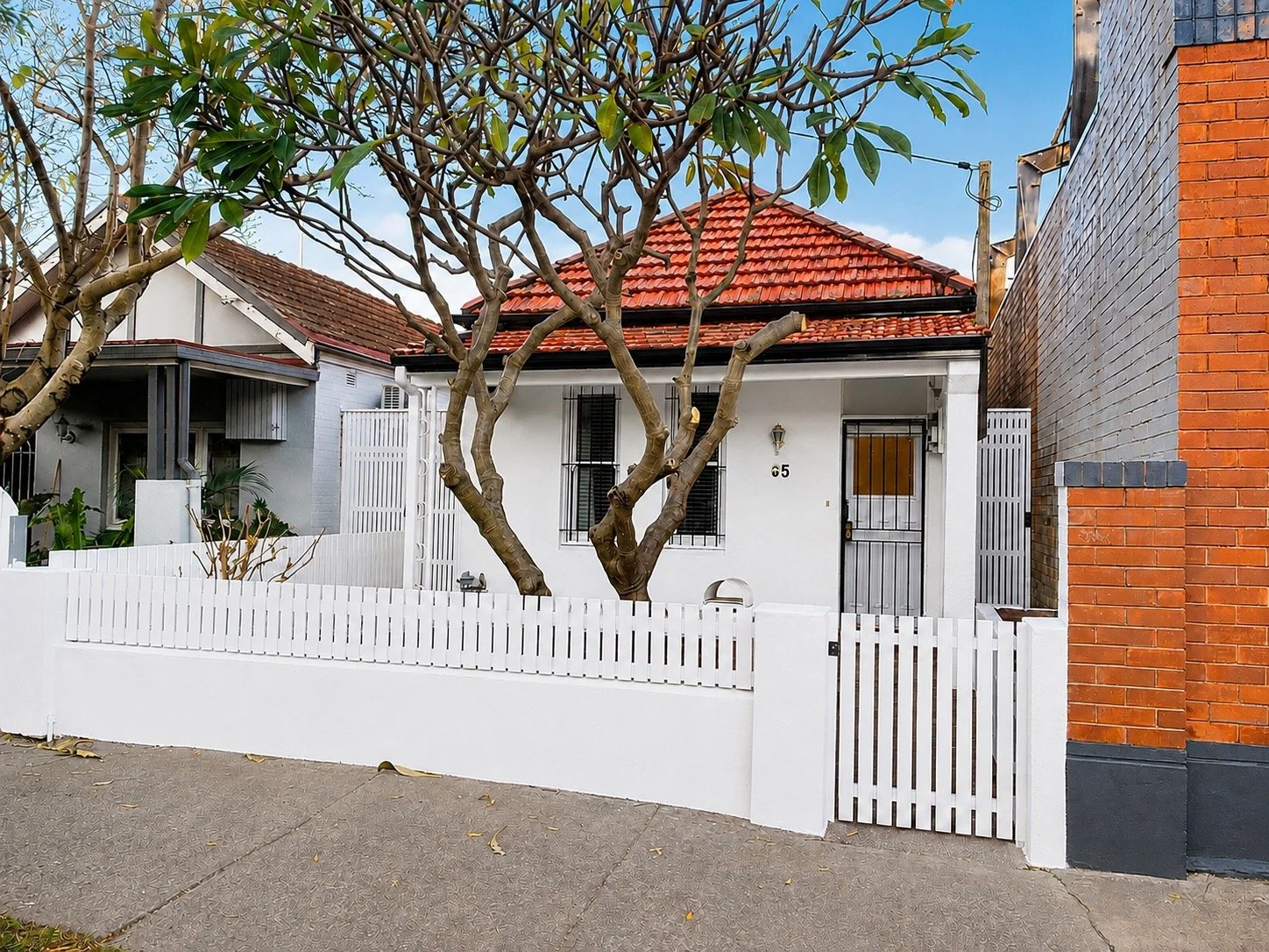 White house in Leichhardt with red-tiled roof, black door, black window bars, surrounded by a white picket fence, on a sidewalk, with a large tree in front, neighboring buildings on each side, and a blue sky with clouds overhead.