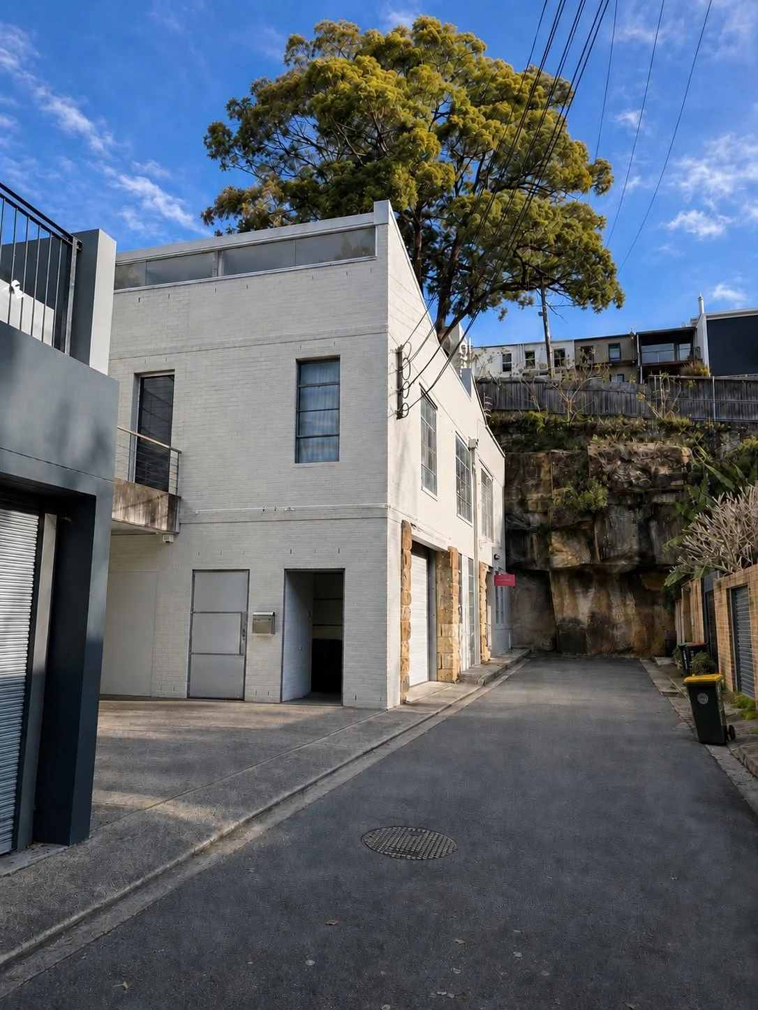 A gallery in Paddington Roslyn Oxley9, white modern multi-story building on a narrow street with a large tree behind it, power lines above, blue sky with some clouds, and rock formations at the back.