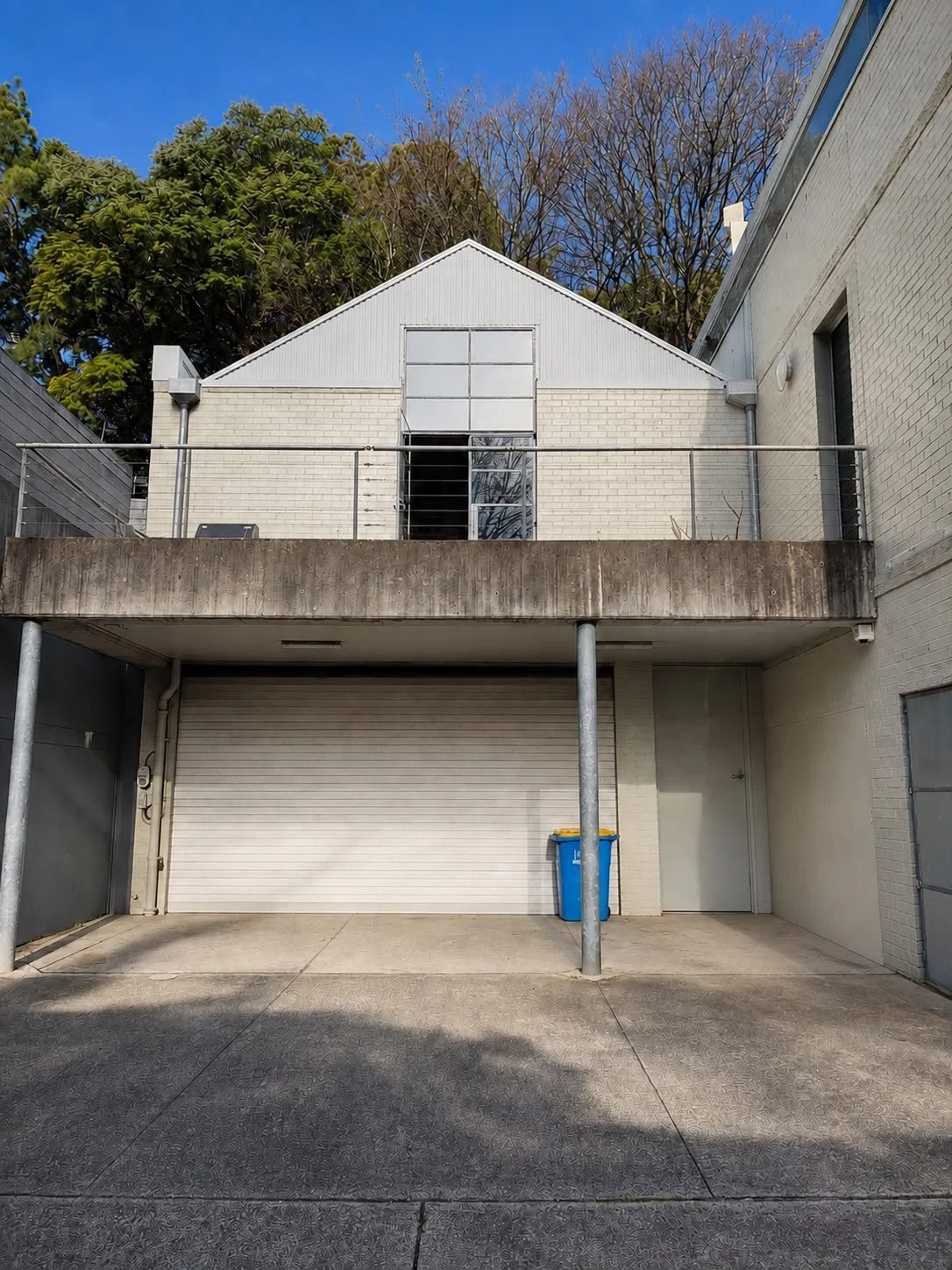 A building with a beige brick exterior, white gabled roof, and a large closed white garage door on the ground level, with a blue recycling bin and a side door nearby. There is an upper balcony with a metal railing, and trees with leafless branches an