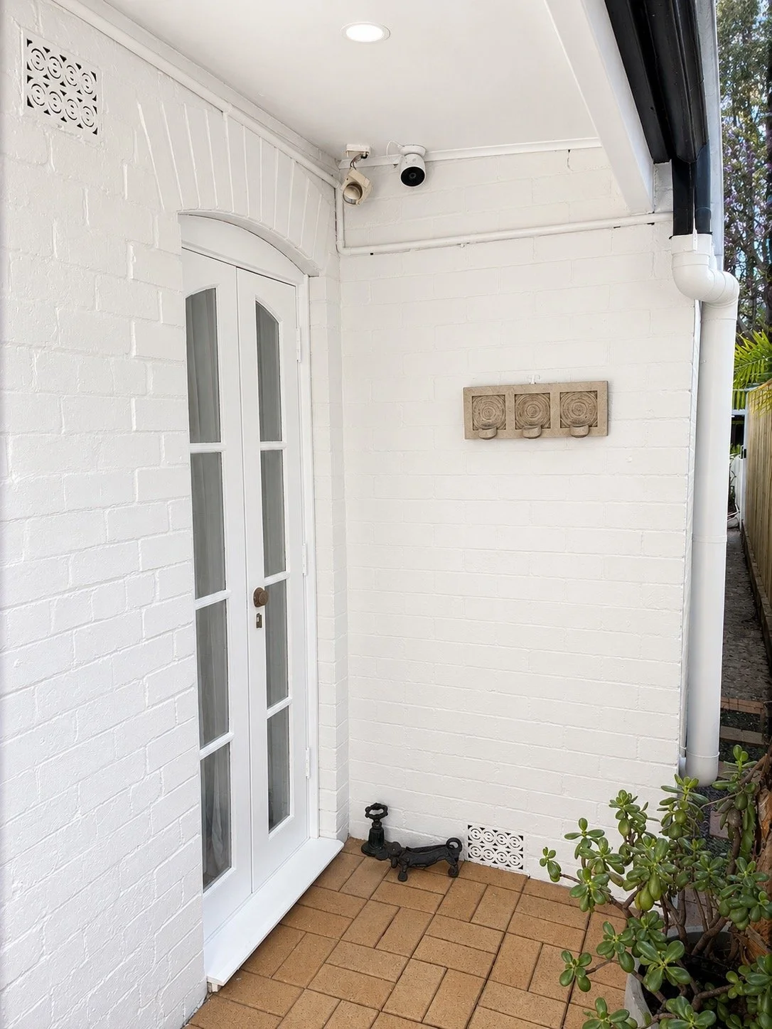 White brick porch with white door, small black security camera, outdoor black faucet on tile floor, decorative wall piece, and potted succulent plant.