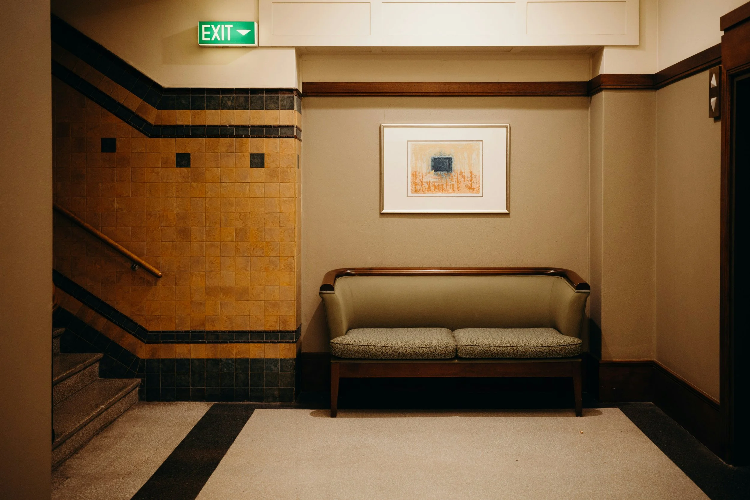 An indoor scene featuring a green exit sign above a beige wall, a framed abstract art piece, a cushioned bench, and a staircase with black and orange tiles.