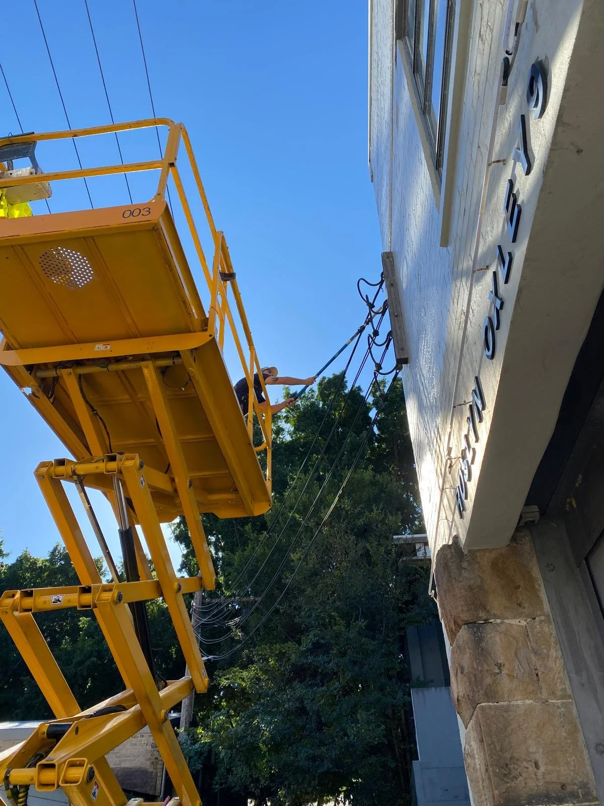 A worker on a yellow aerial lift cleaning or inspecting a large outdoor sign with engraved letters. The sign is mounted on a stone base and the sky is clear with trees in the background.