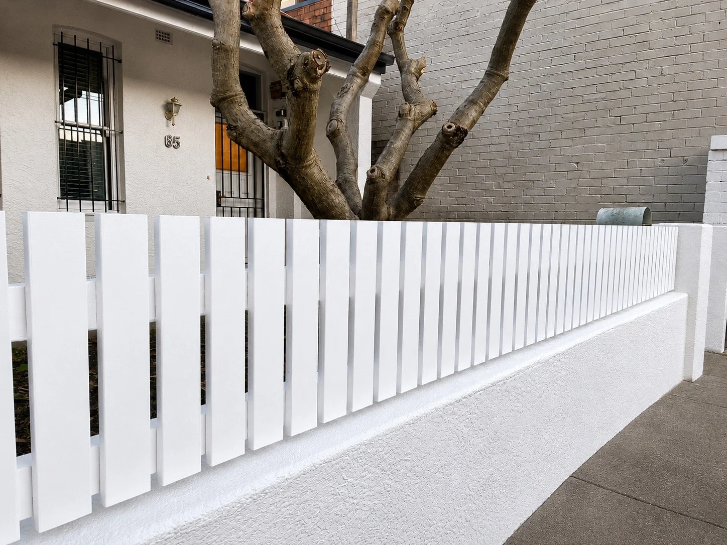 A front view of a white wooden fence in front of a house in Leichhardt with a tree behind the fence and two windows with black bars. There is a house number 85 on the wall of the house, and a cylindrical object on the right side of the fence.