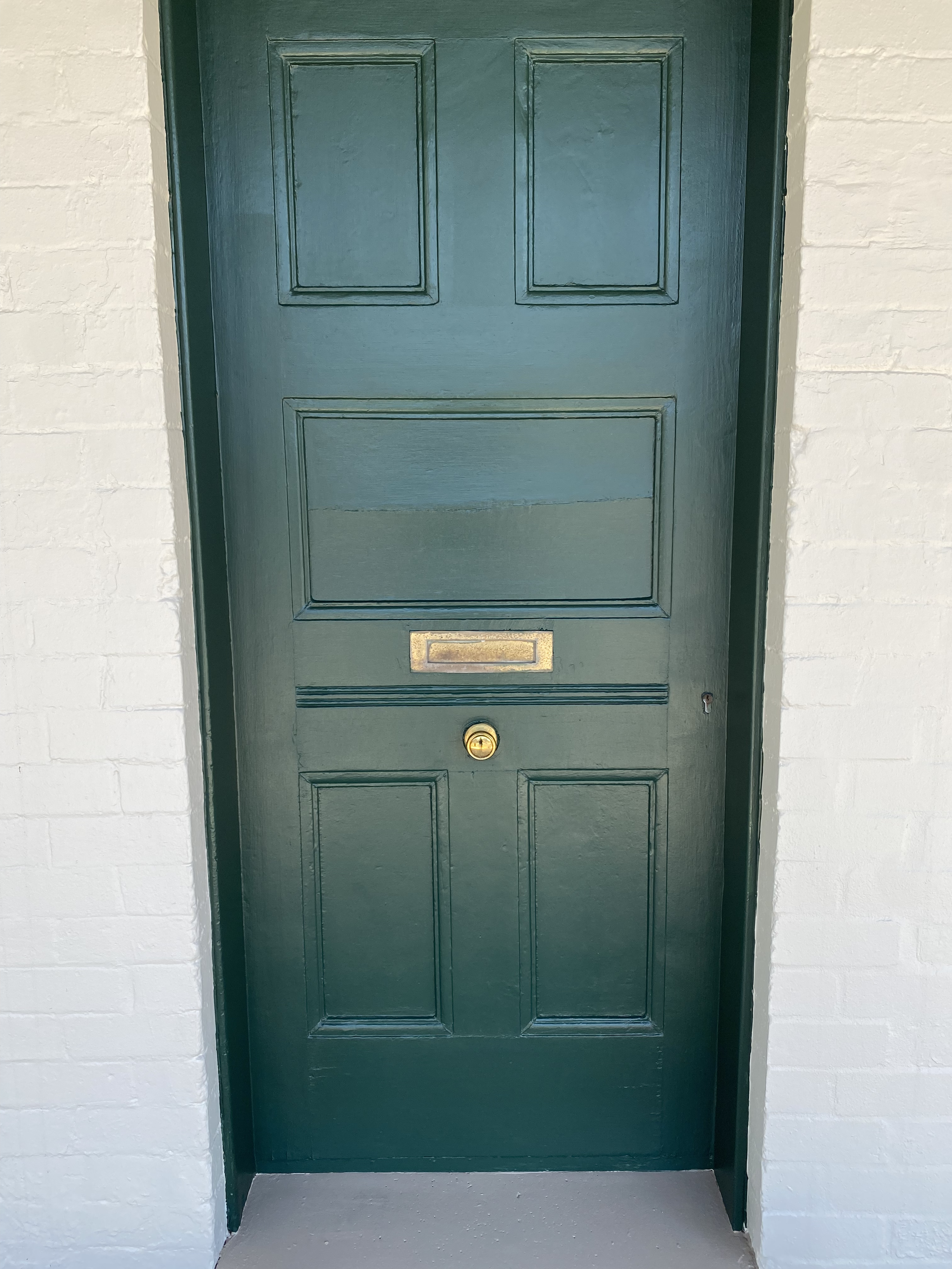Green front door with a mail slot, peephole, and surrounding white brick wall.